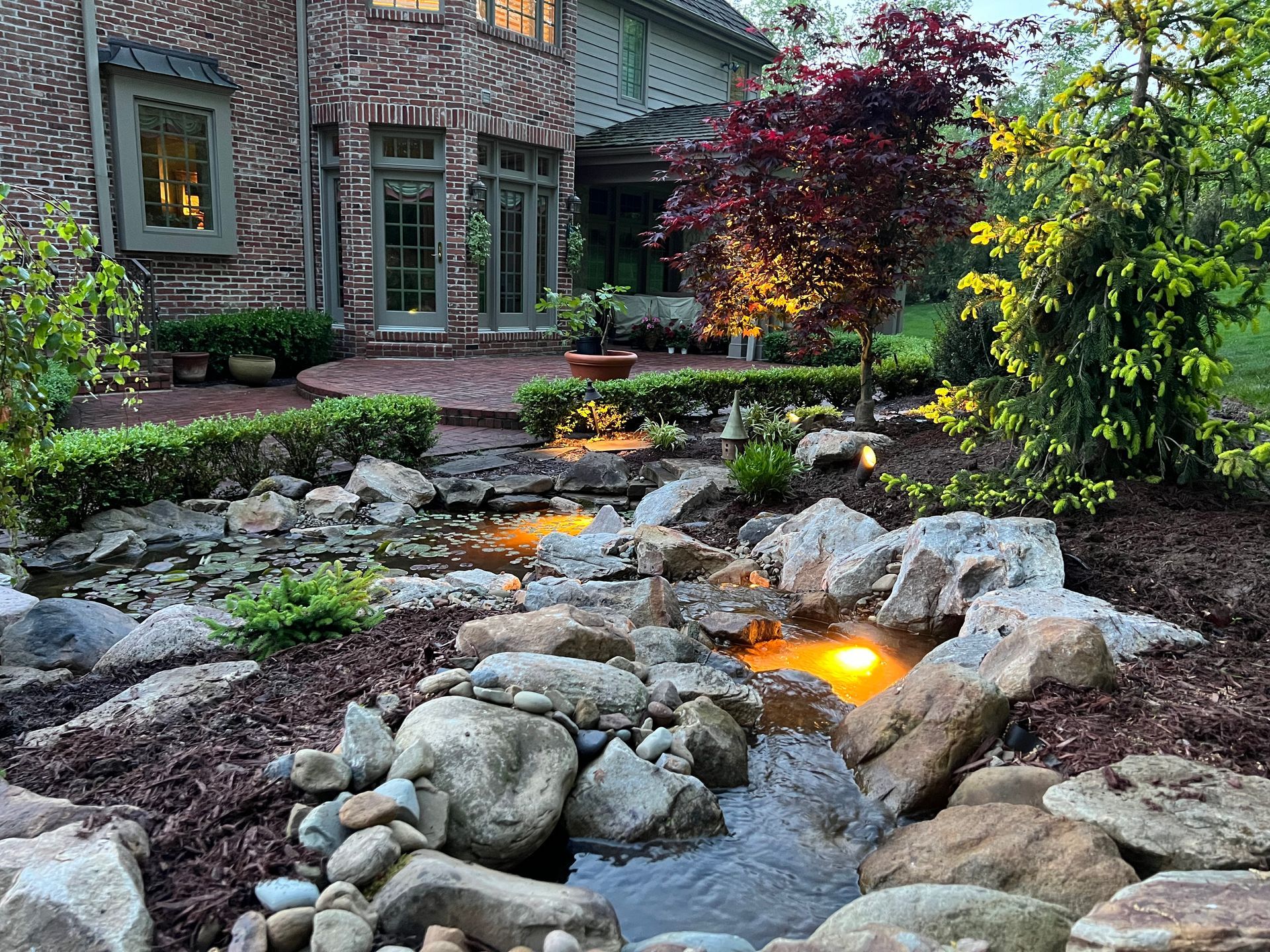 Illuminated backyard stream with rocks, landscaped garden, and a house patio at dusk