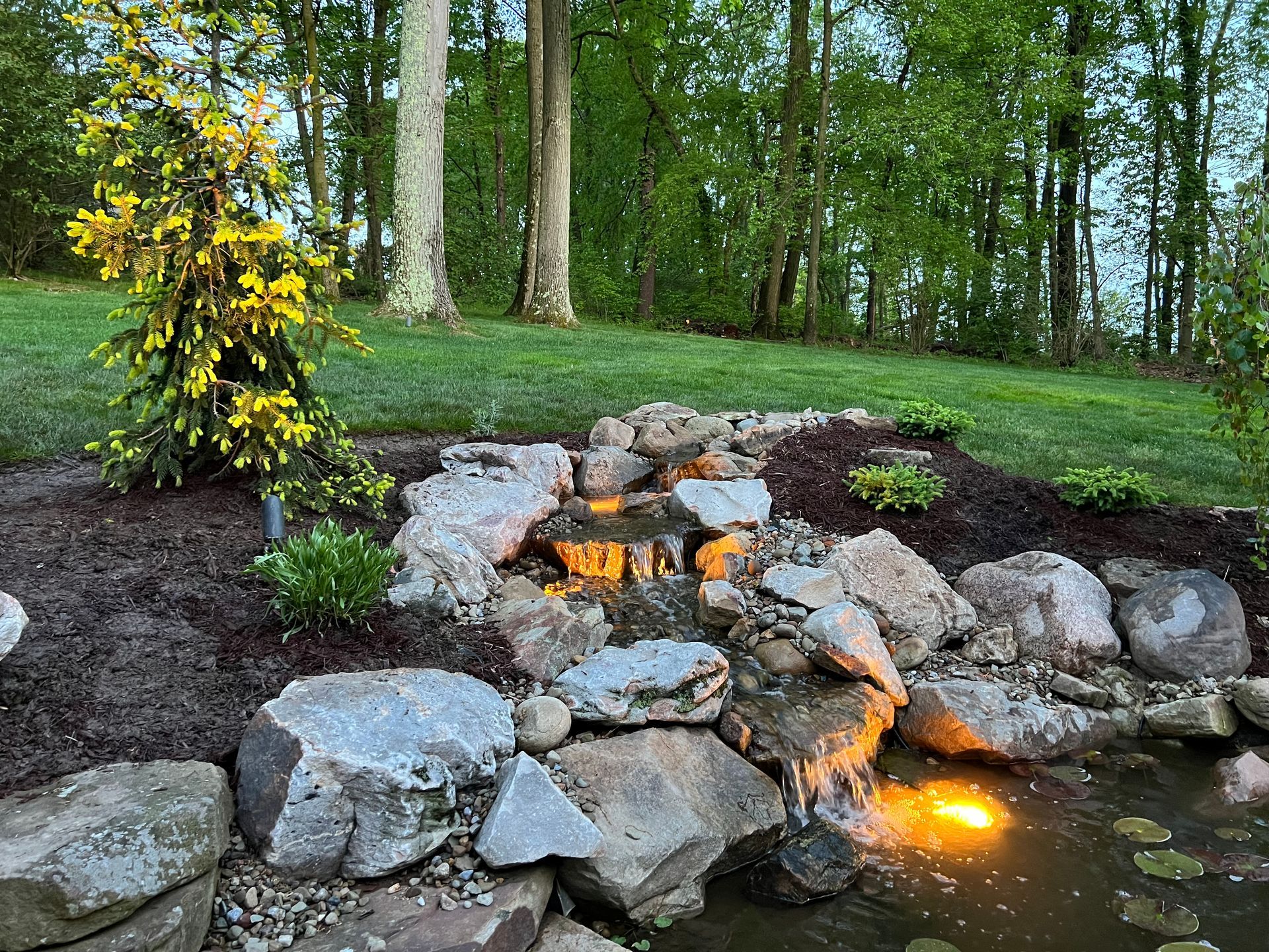 Small backyard stone waterfall with glowing lights, surrounded by rocks and greenery