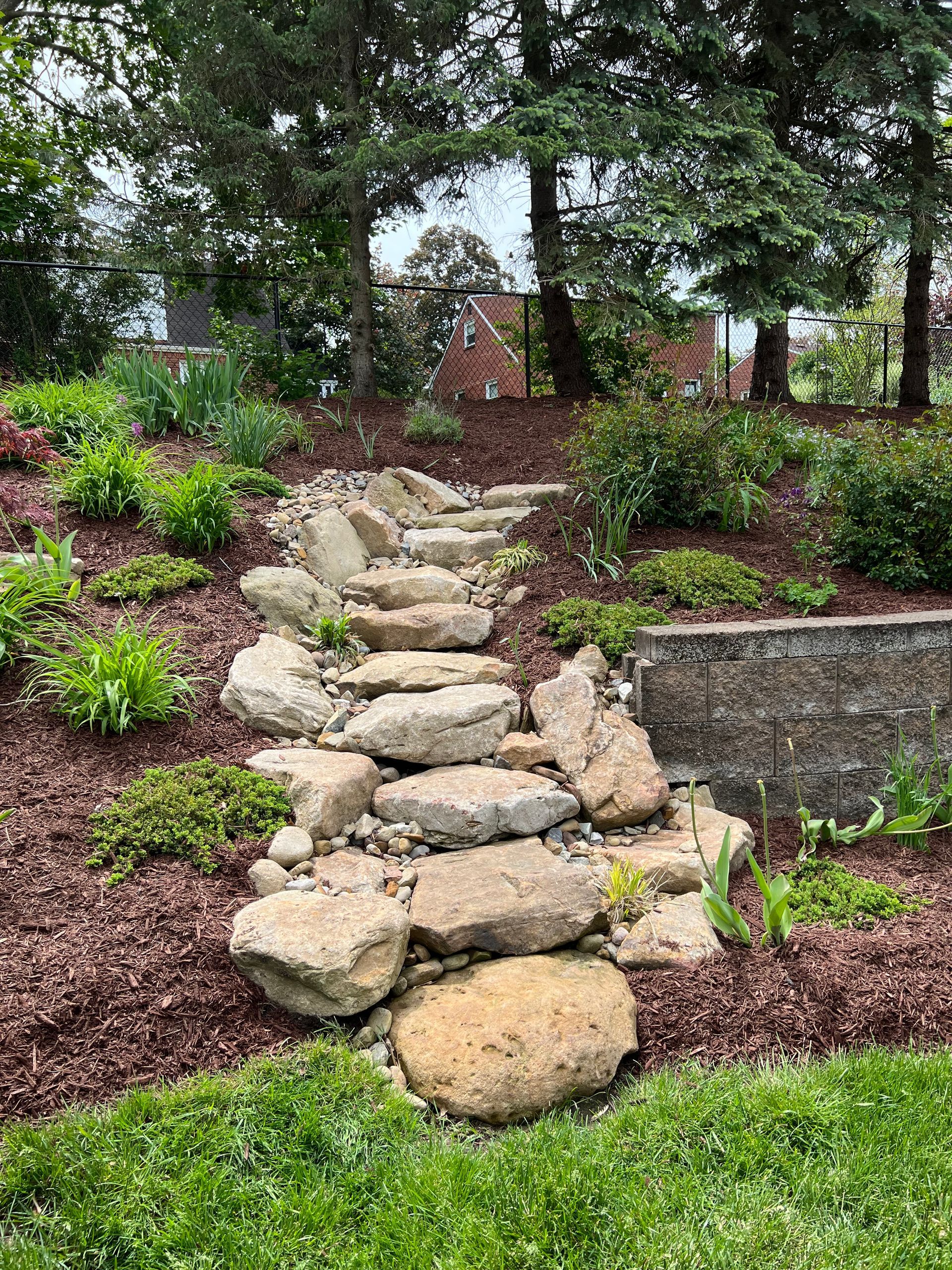 Stone path steps through a lush garden with trees and pink flowers leading uphill to a wooden structure.