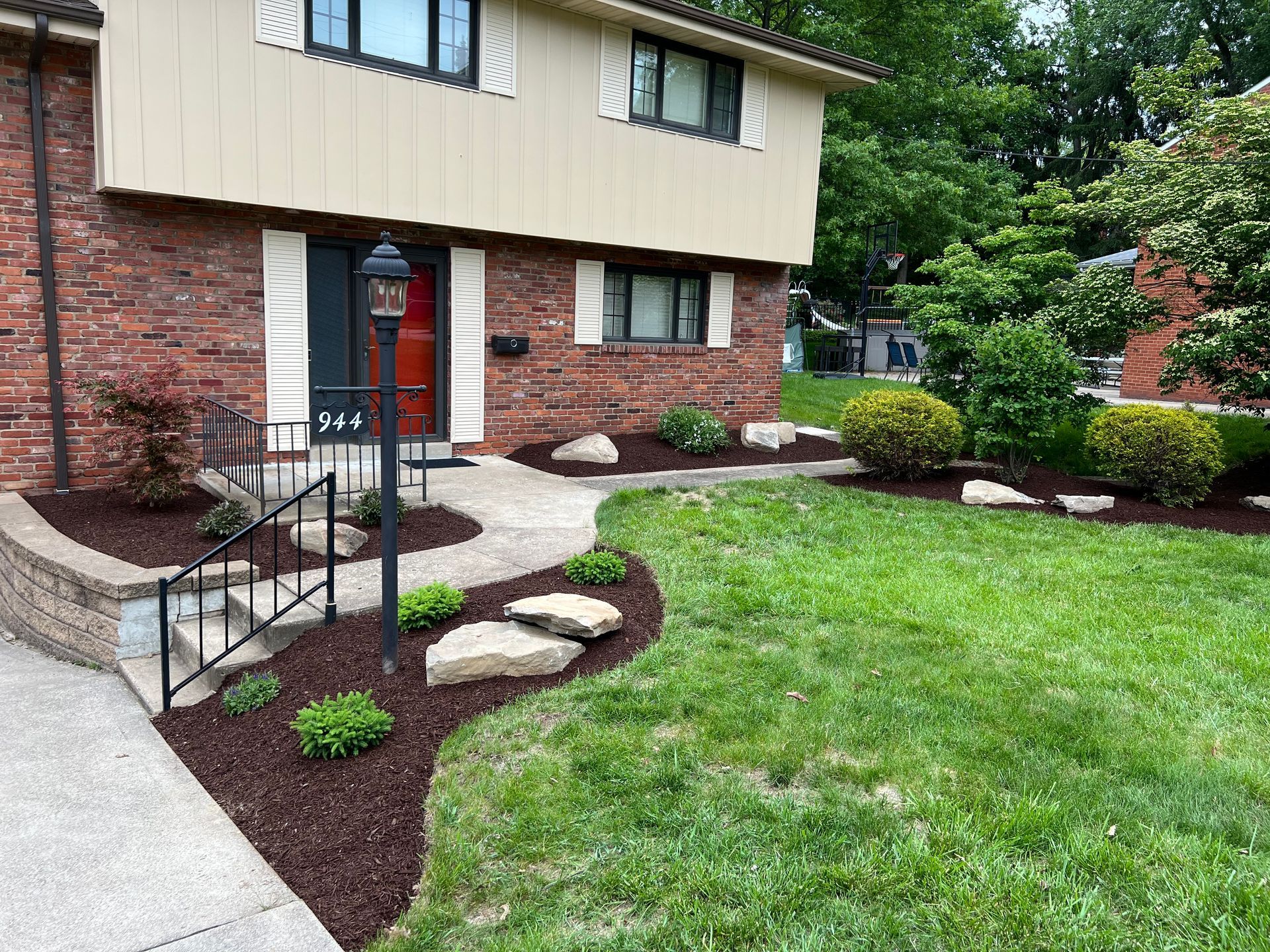 Suburban house entrance with brick and tan siding, front steps, and landscaped lawn with mulch beds