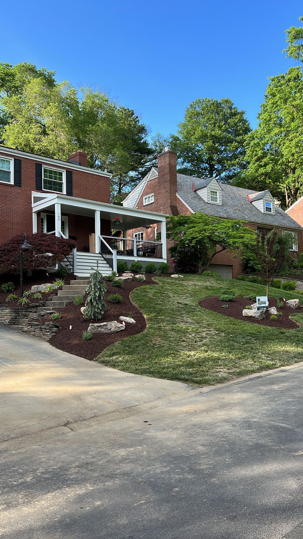 Brick house with white porch, landscaped front yard, and driveway under a blue sky