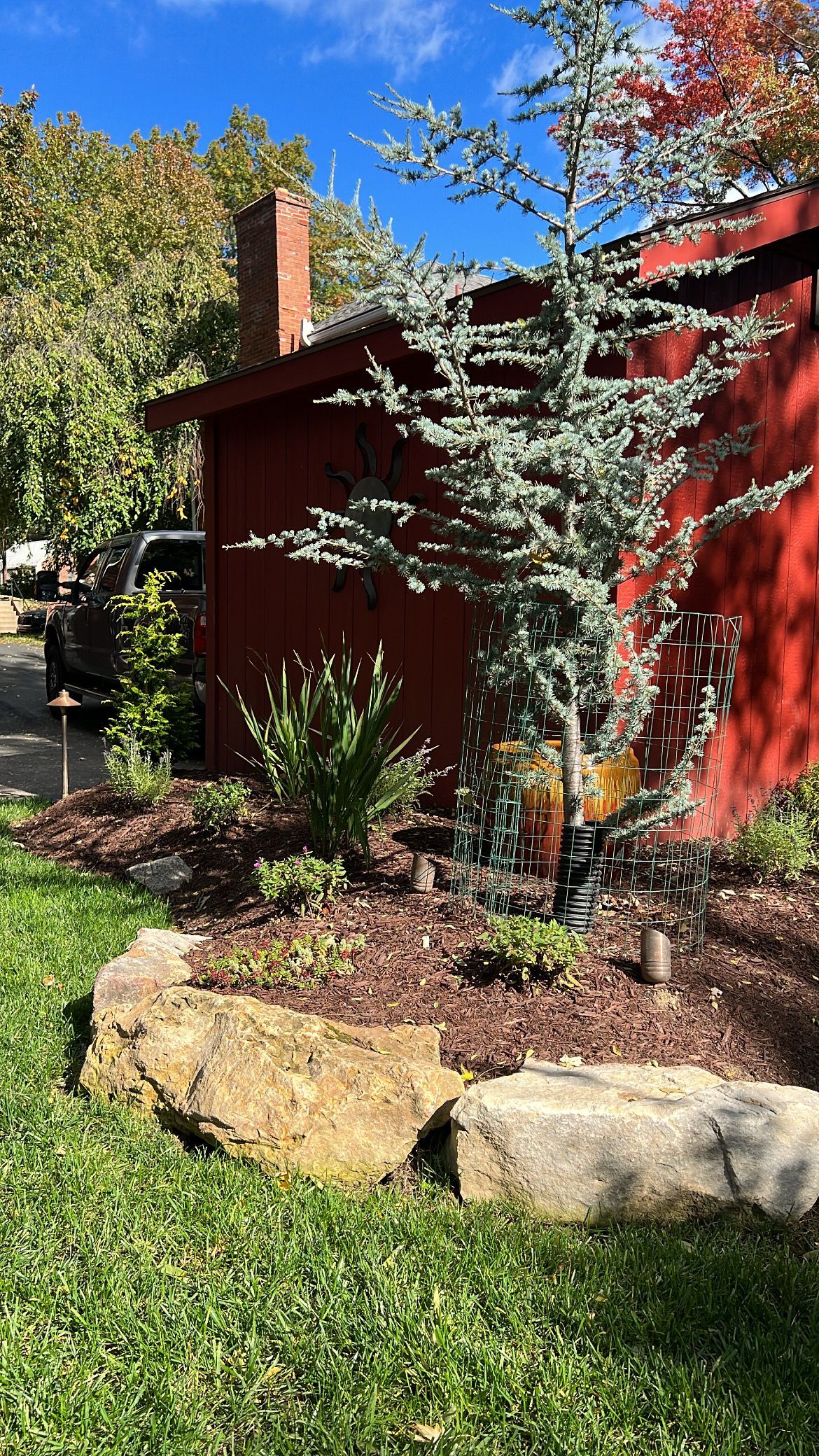 Red house with chimney, framed by trees and shrubs, beside a stone-edged garden bed.