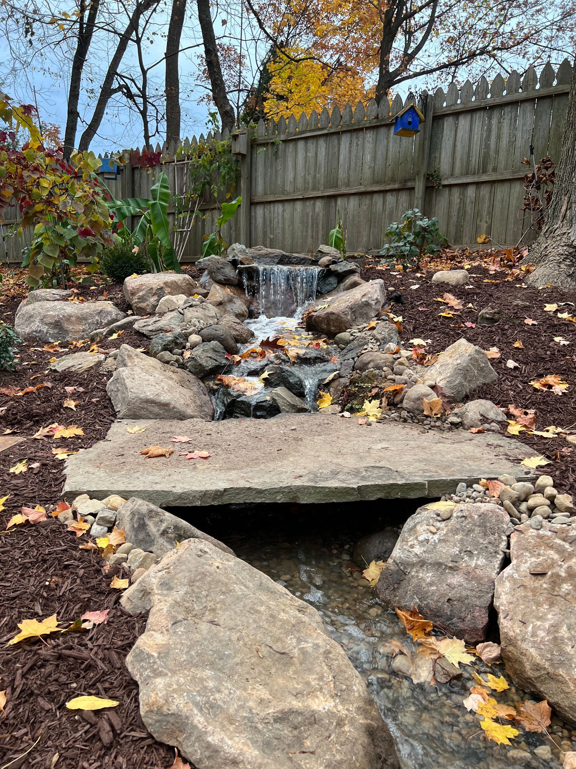 Rock garden waterfall with a small stream flowing over rocks behind a wooden fence in autumn.