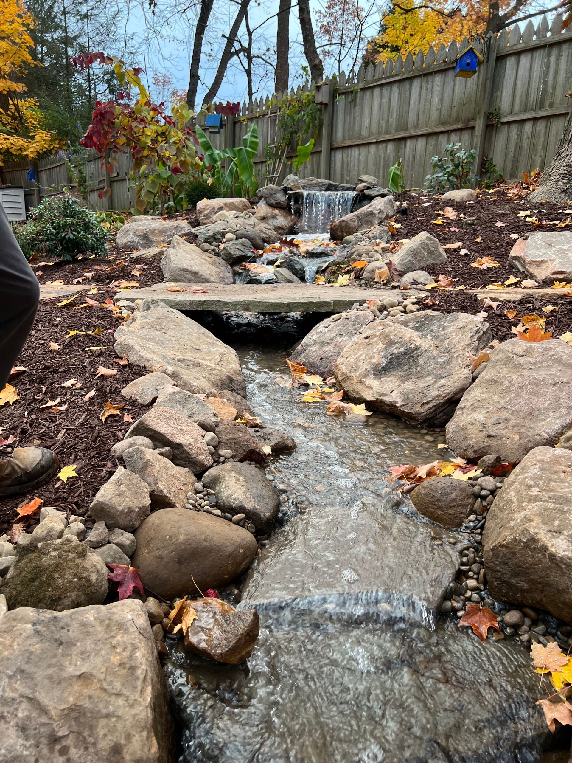 Rock-lined backyard stream with small waterfall and autumn foliage by a wooden fence