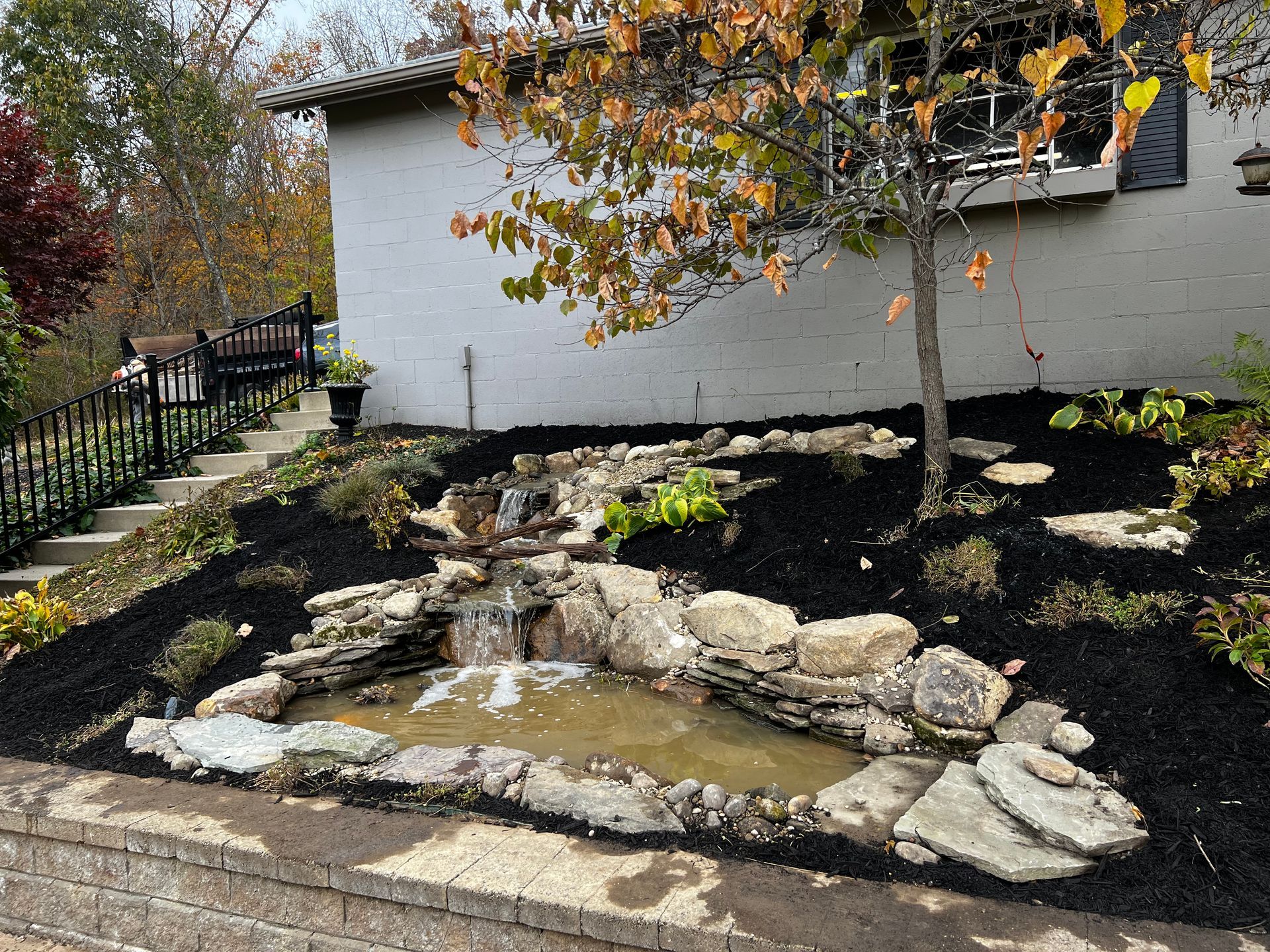 Stone-lined garden pond beside a white house with autumn trees and landscaping