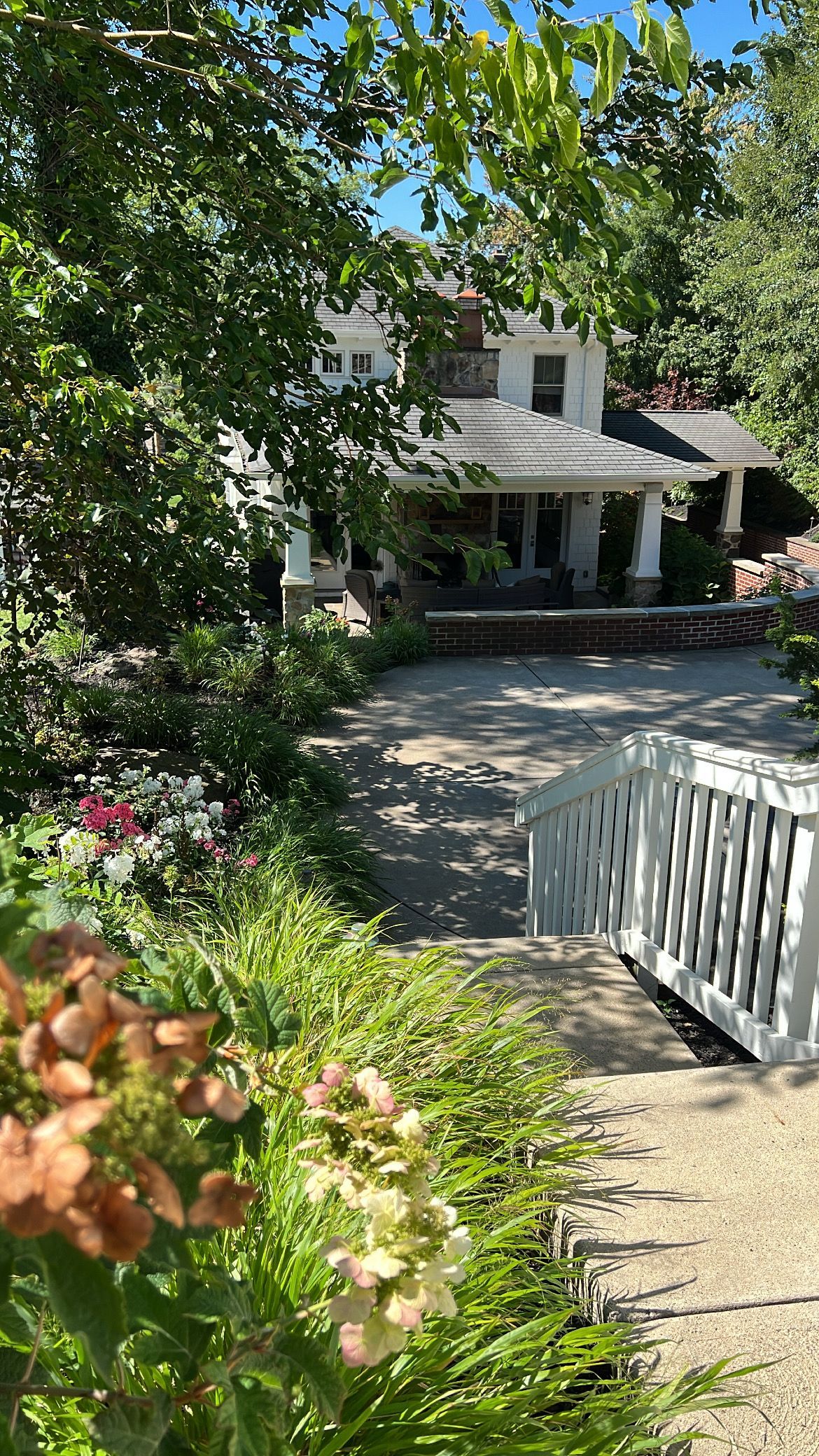White house with porch behind a garden path and white fence, shaded by leafy trees.