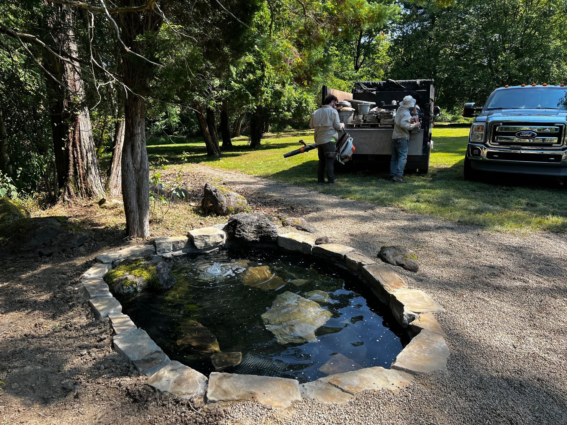 Two people standing by a pickup truck near a small stone-lined pond in a wooded park area