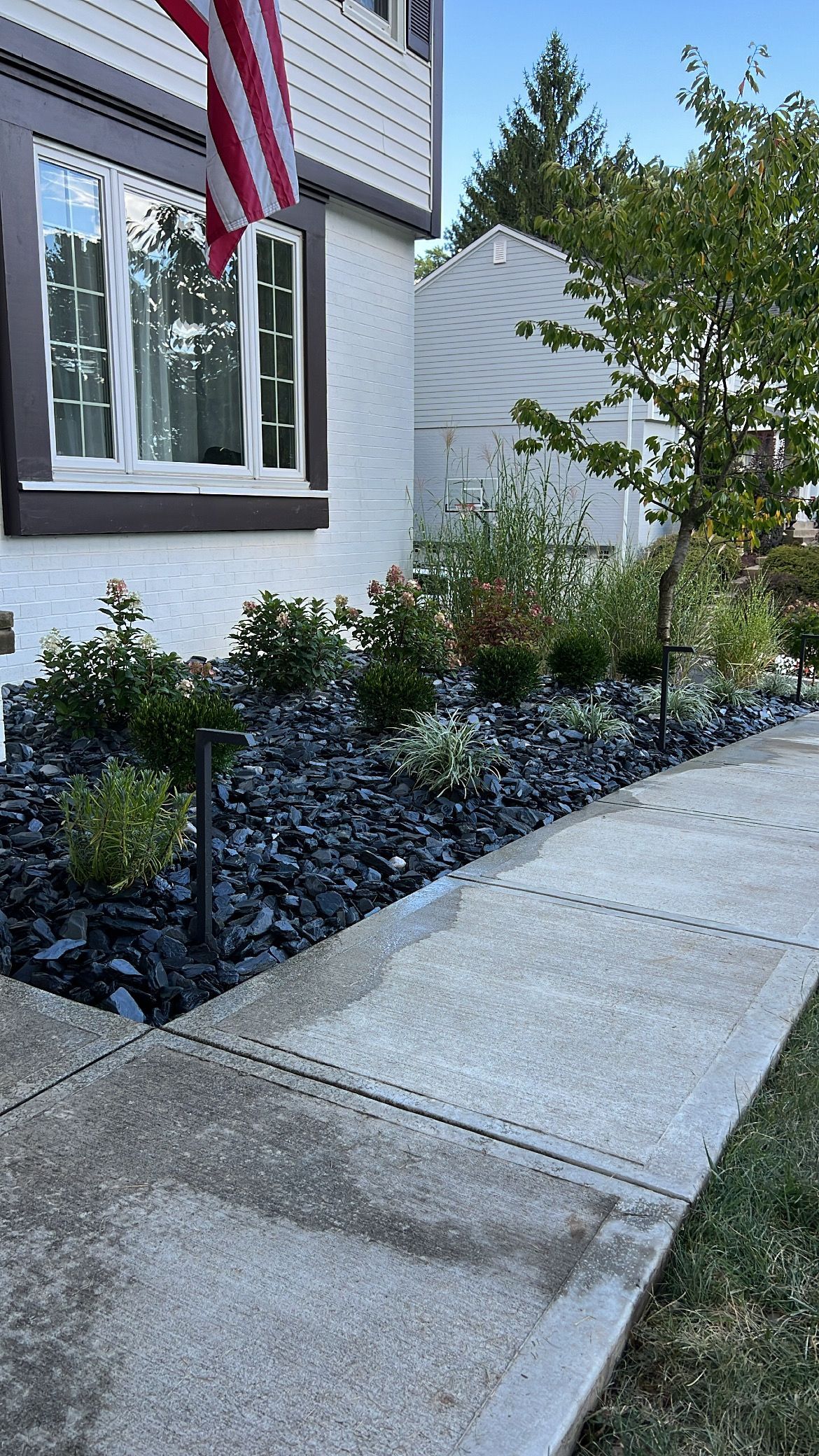 Front walkway beside a house with an American flag, stone path, and landscaped garden beds under a blue sky