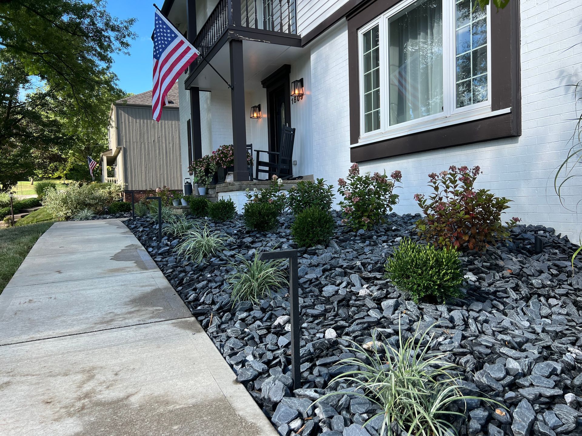 Front walkway beside a white house with an American flag and a landscaped rock garden.