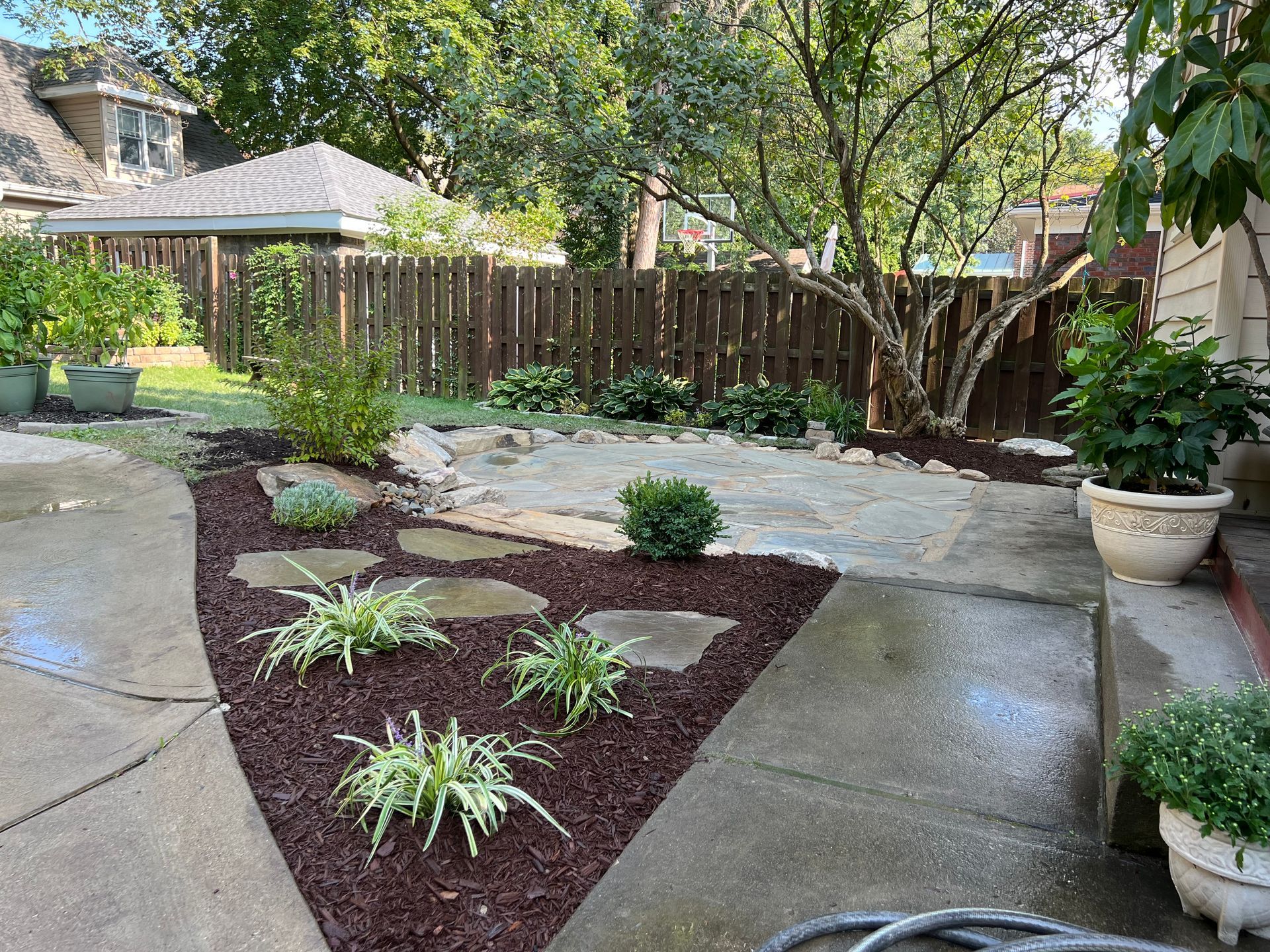Landscaped backyard patio with concrete paths, mulch bed, potted plants, and a wooden fence.