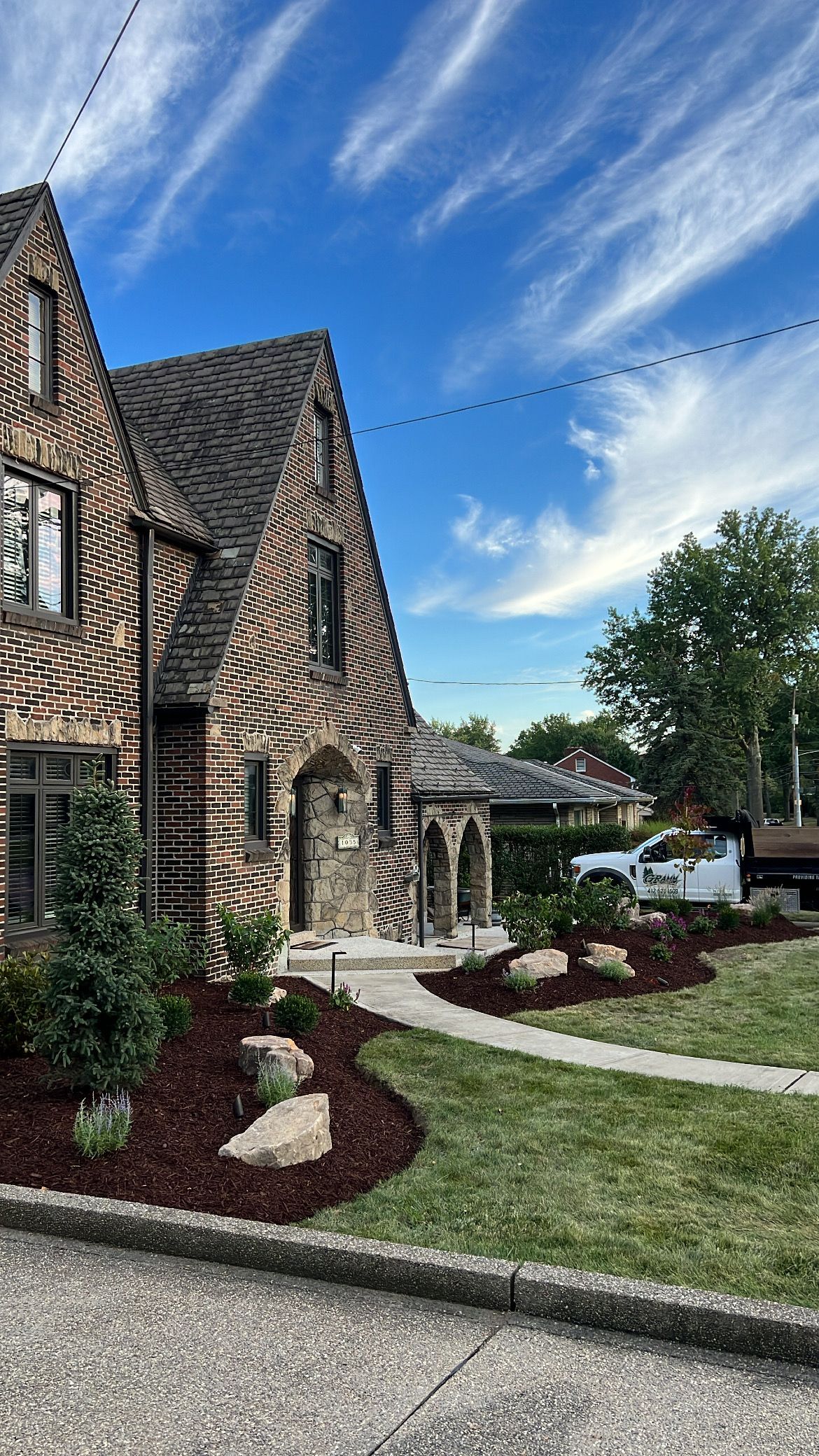 Stone house with steep gabled roof, landscaped flower beds, and a blue sky with wispy clouds