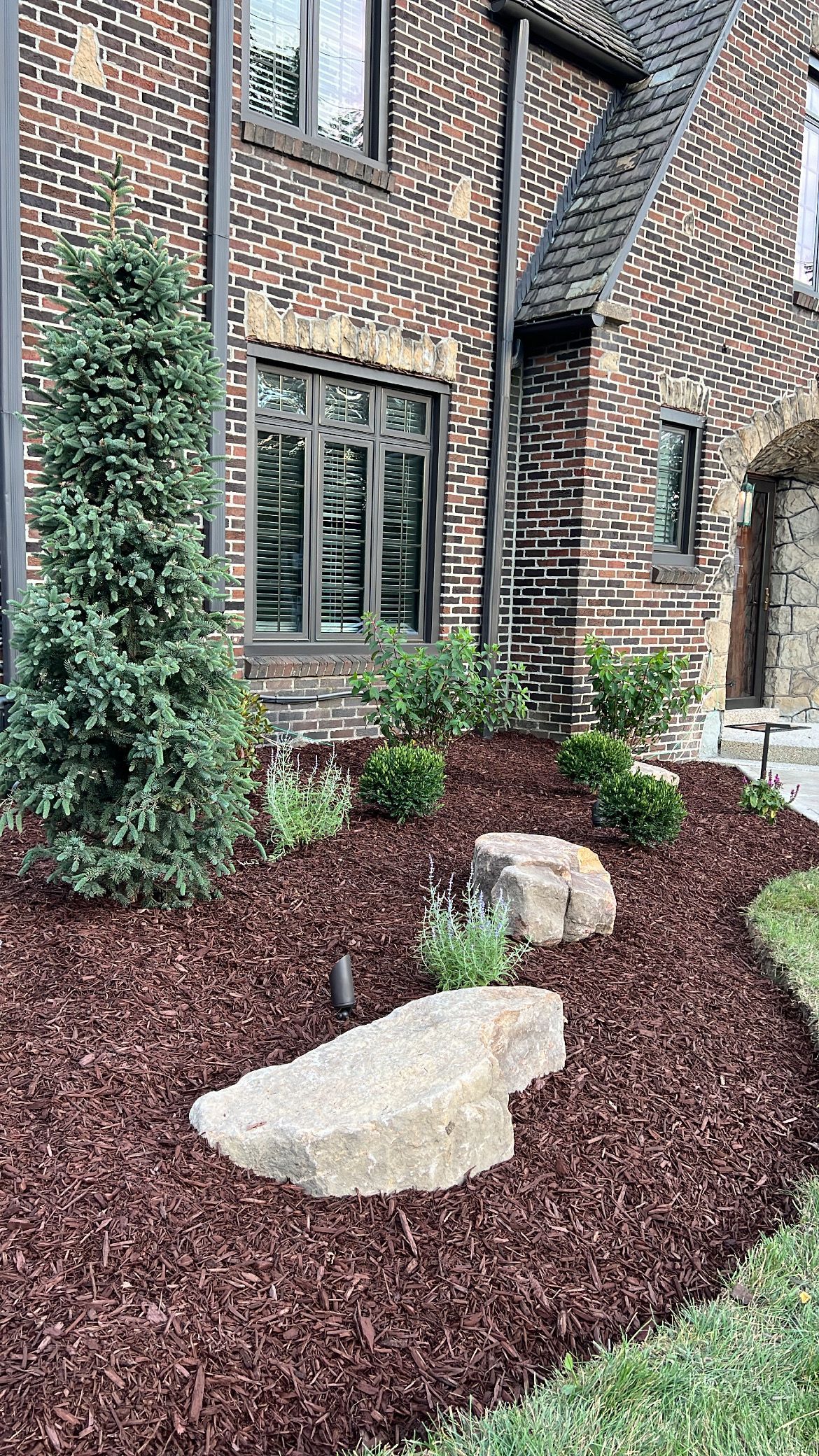 Brick townhouse exterior with windows, leafy shrubs, and a landscaped bed with large rocks