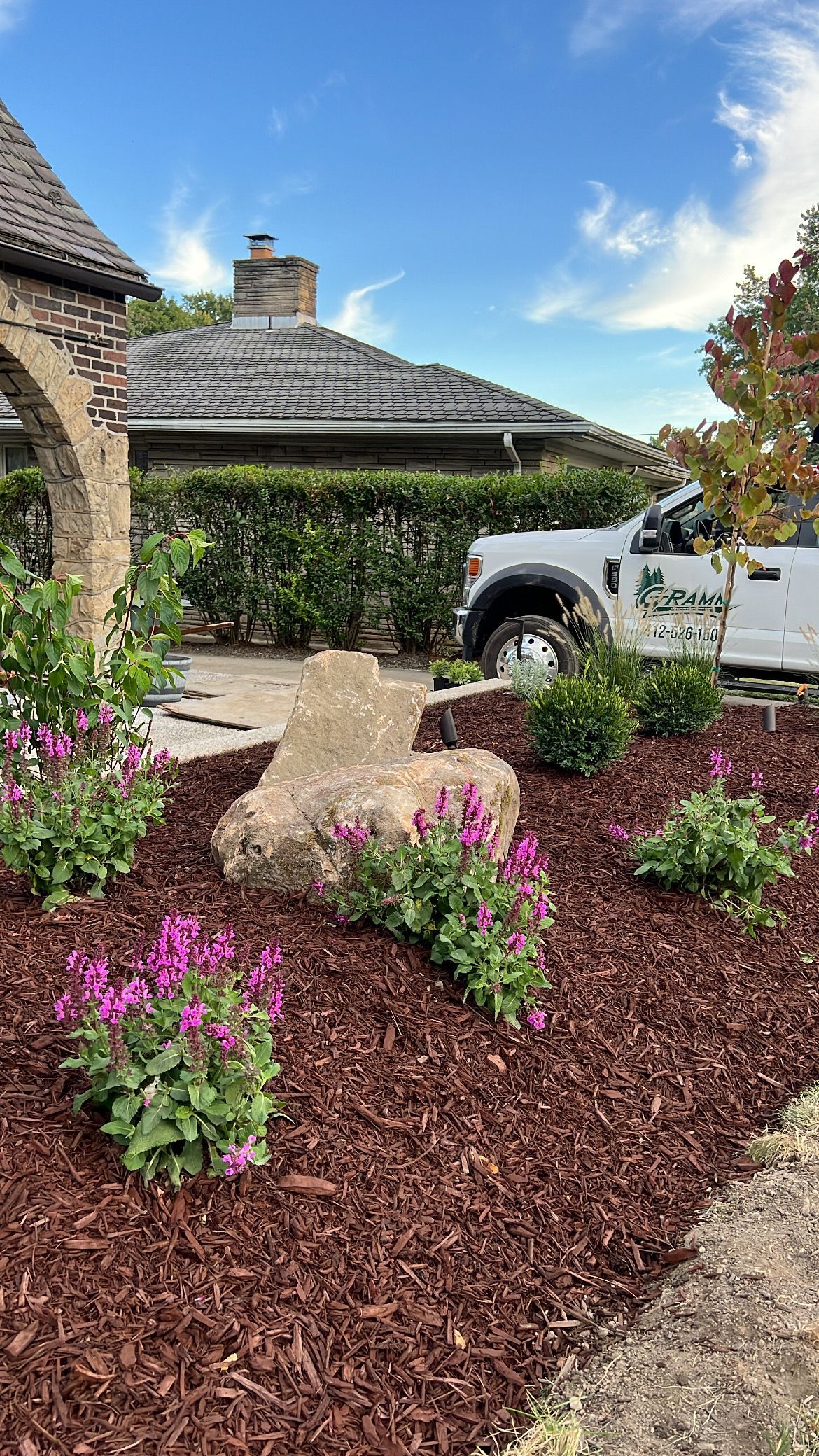 Landscaped front yard with pink flowers, mulch beds, stone path, and a parked white truck near a house