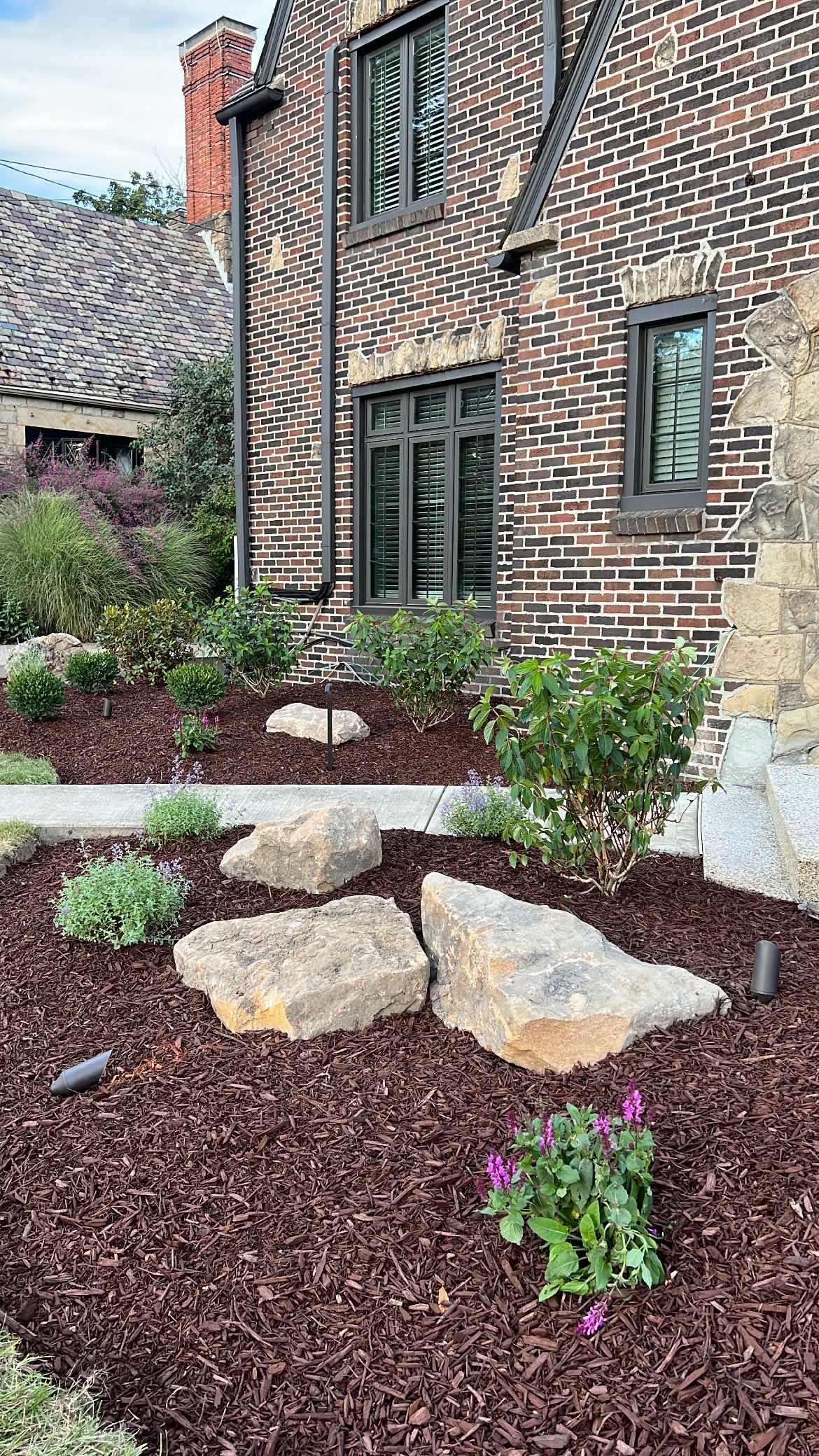 Brick house exterior with landscaped front garden, mulch, rocks, and green shrubs.