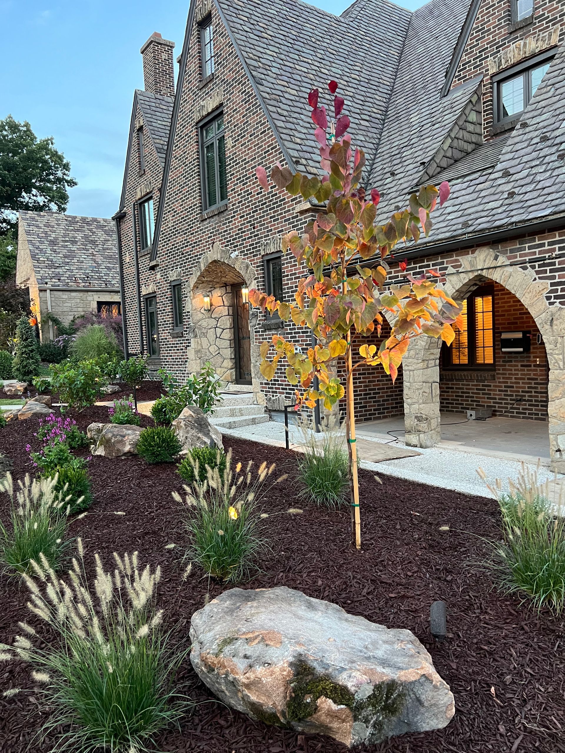 Stone building with arched entryway, landscaped garden, and a small autumn tree in front
