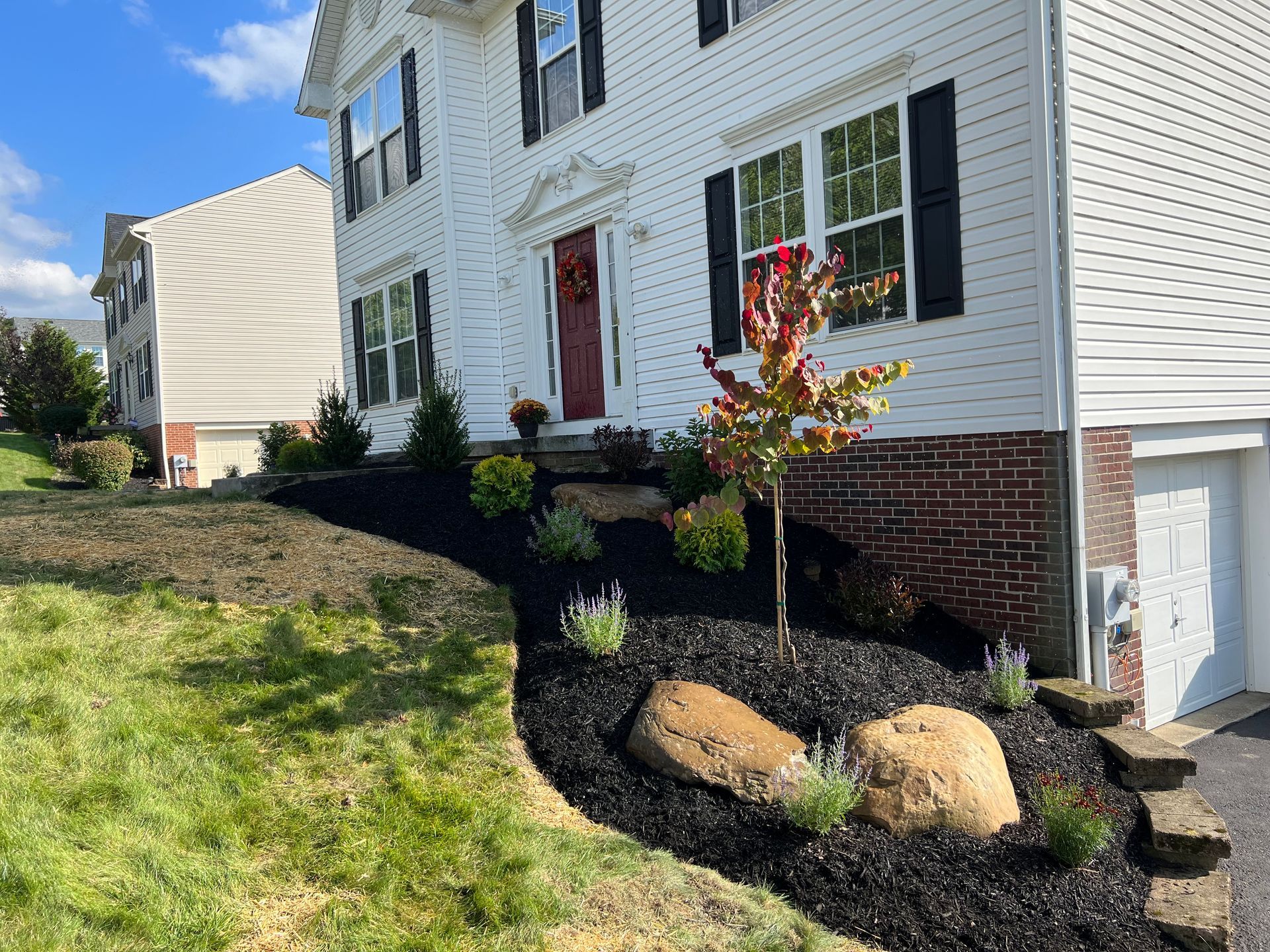 White house exterior with black shutters, brick foundation, and landscaped flower bed on a sloped lawn