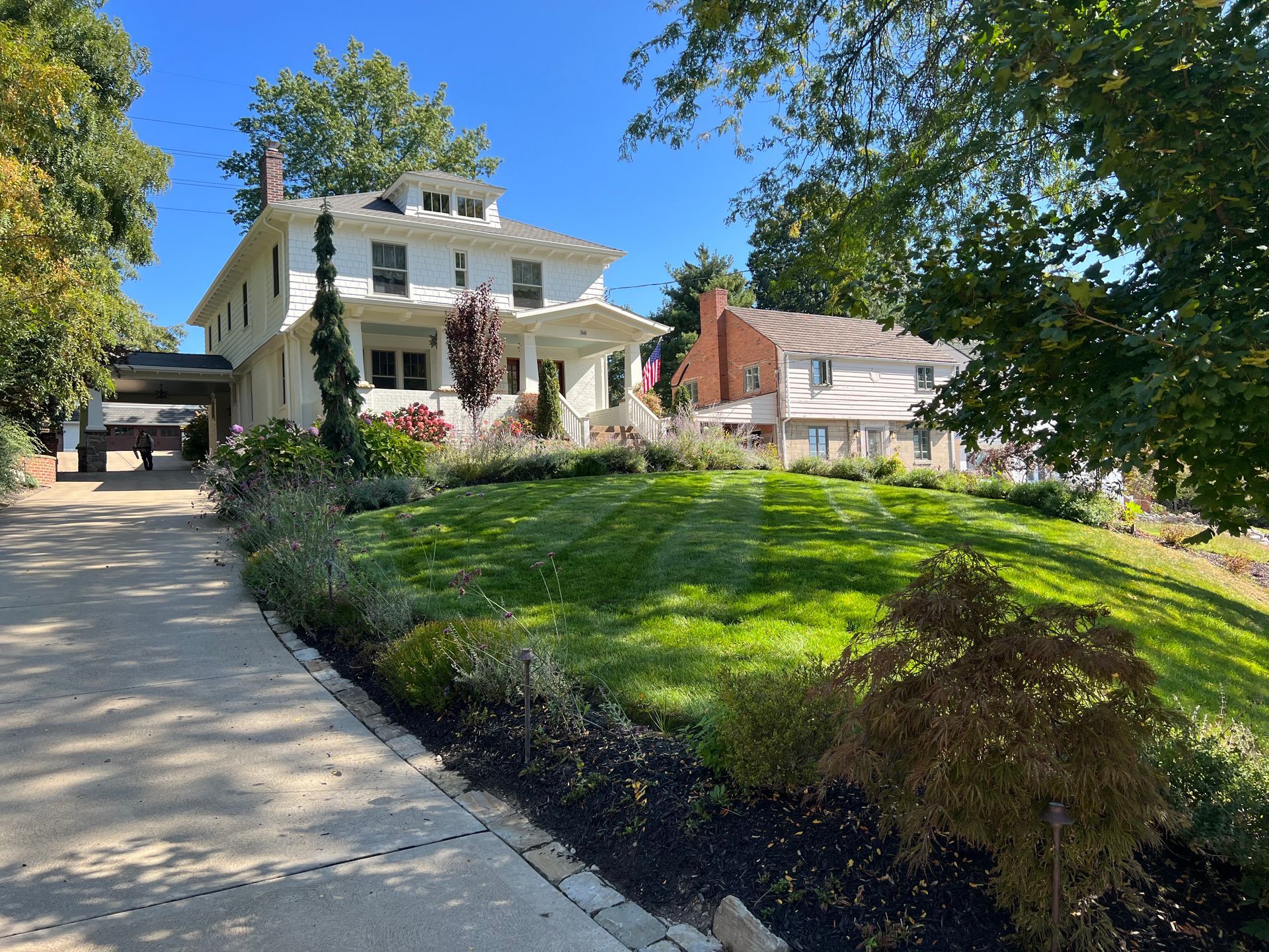 Large white house with a porch on a grassy hillside, viewed from a driveway under blue sky