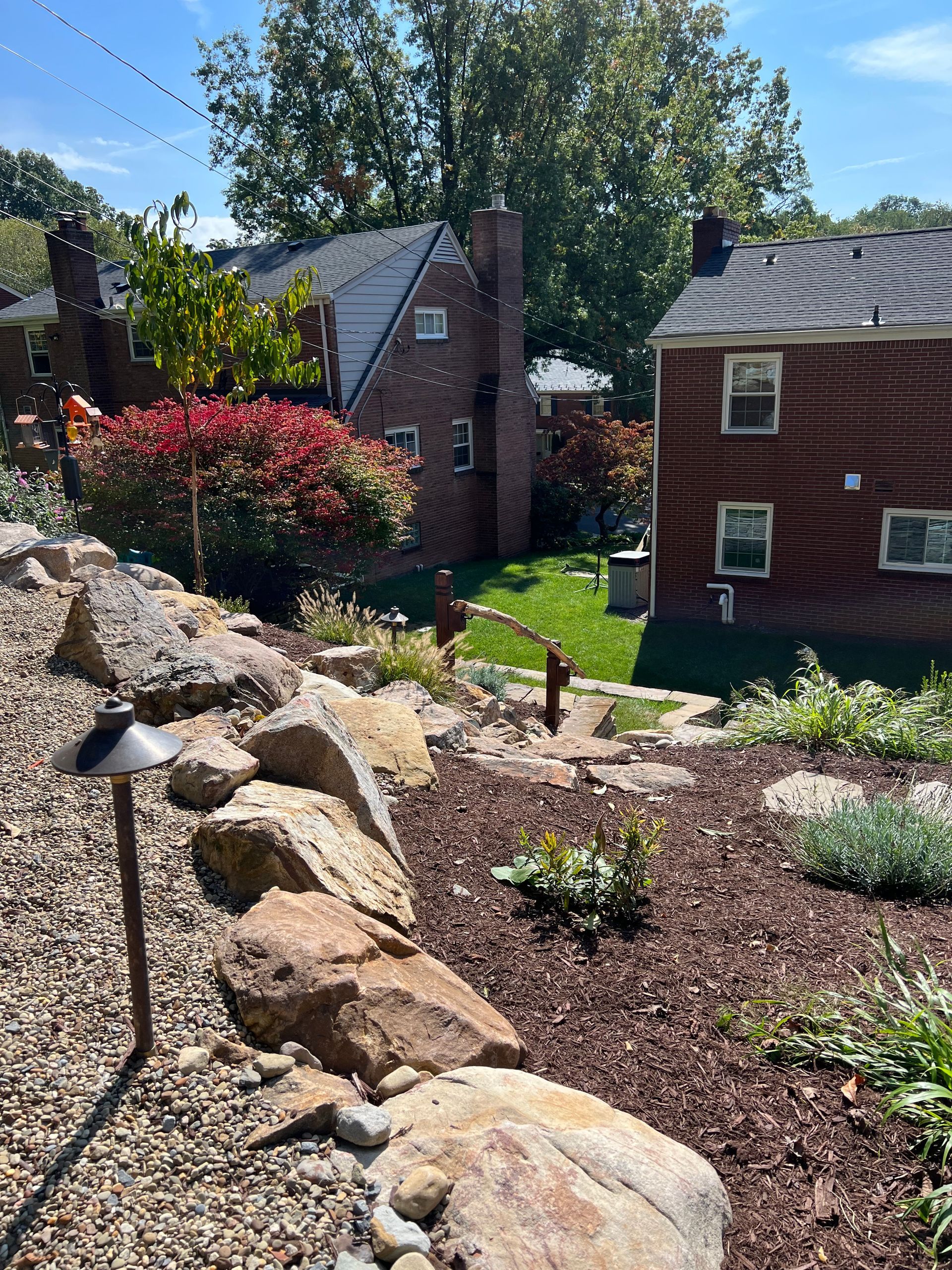 Rock garden with large boulders, mulch, and small plants beside brick houses on a sunny day