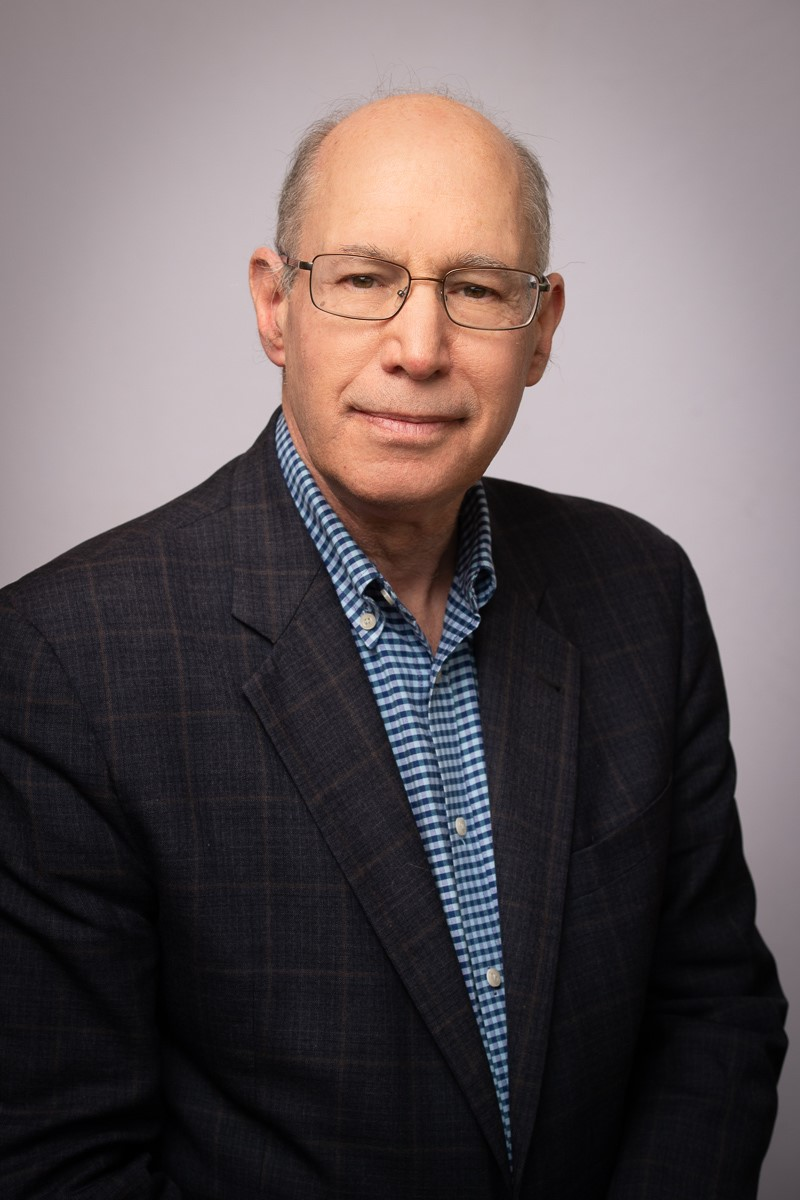 Portrait of a man in glasses wearing a dark blazer and blue checkered shirt against a gray background