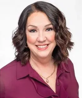 a woman in a purple shirt and necklace is smiling for the camera .