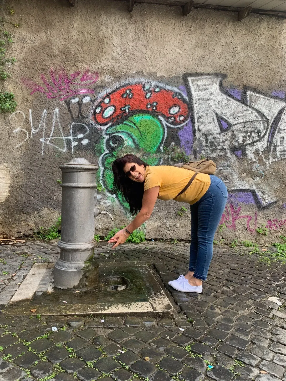 a woman is drinking water from a fountain in front of a graffiti covered wall .