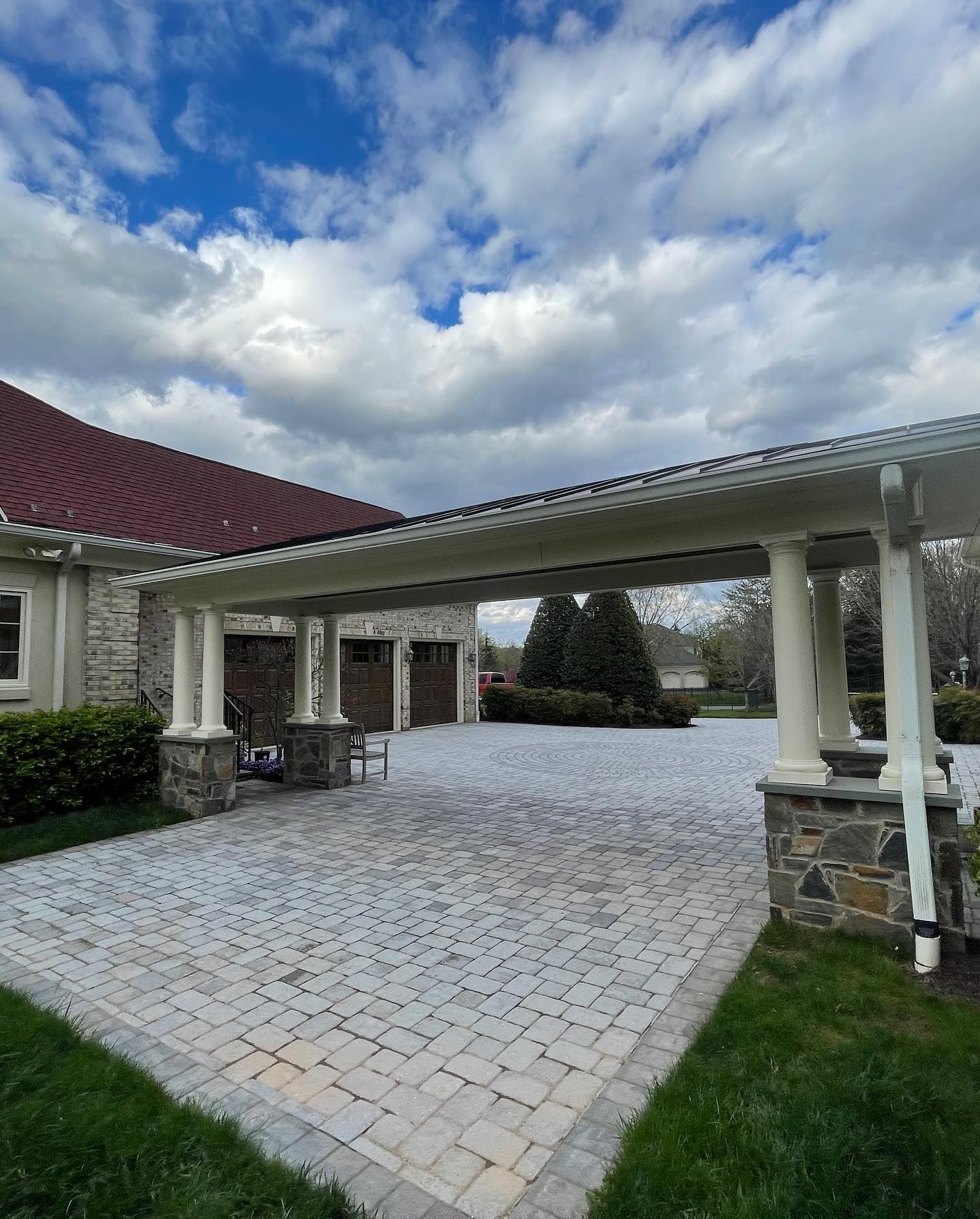 A covered walkway with white columns leads to a house with a red roof, stone walls, and a light-colored stone paver driveway.