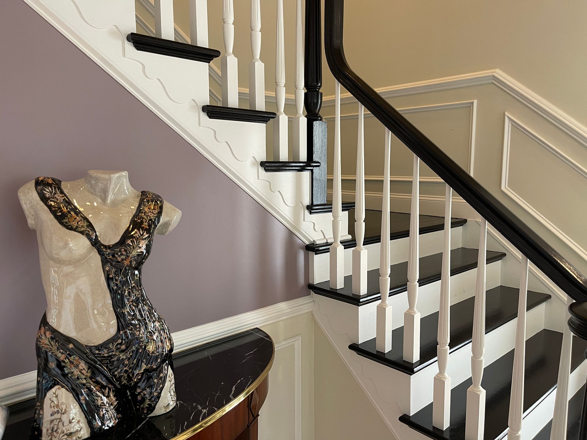 Elegant interior staircase with black treads, white balusters, and dark wood handrail, beside a decorative ceramic torso sculpture on a marble-top table against a mauve and white paneled wall.