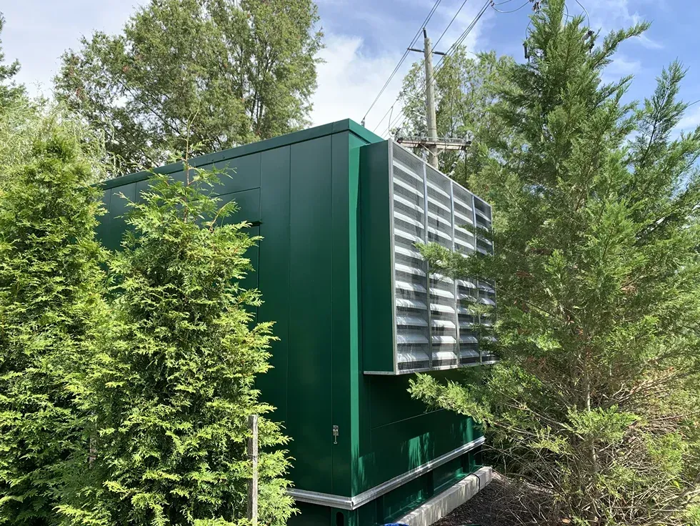A green utility enclosure with a louvered ventilation panel, surrounded by pine trees under a blue sky.