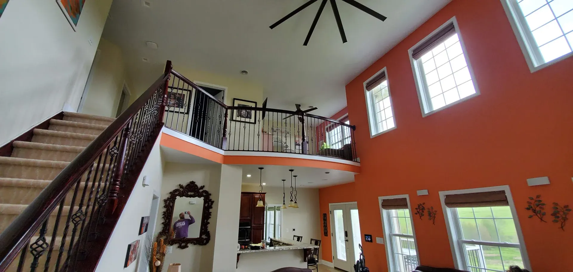A bright foyer with a staircase, a balcony overlooking the kitchen, and orange walls with high windows.