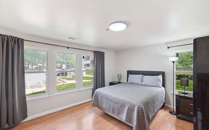 A bedroom with a grey bed, dark curtains, light wood floors, and a floor lamp near the window.