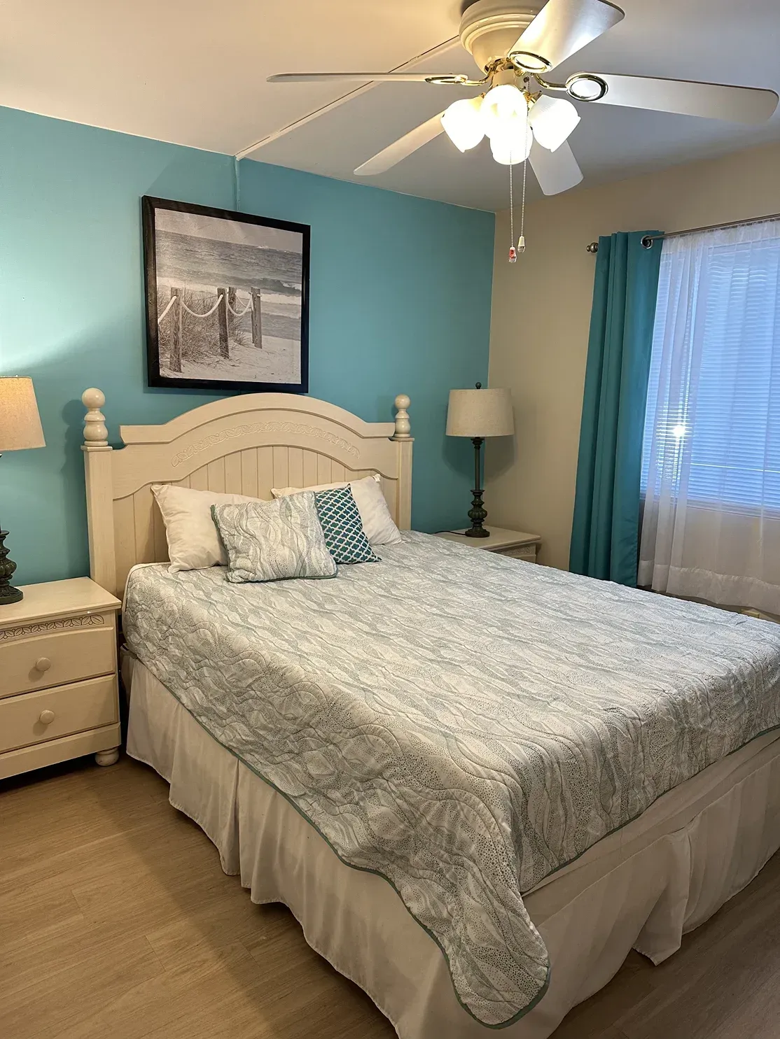 A bedroom with a bed featuring a white headboard, blue-patterned quilt, and a framed beach photo on a turquoise wall.