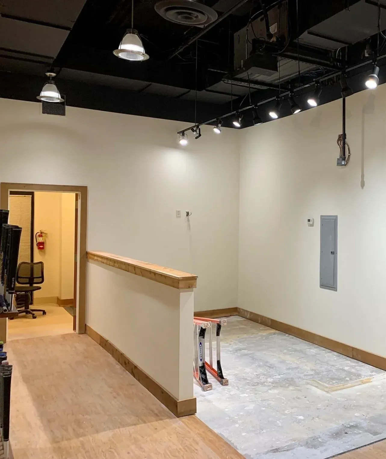 An unfinished commercial room with white walls, track lighting, light-colored flooring, a partial wall, and an open doorway.