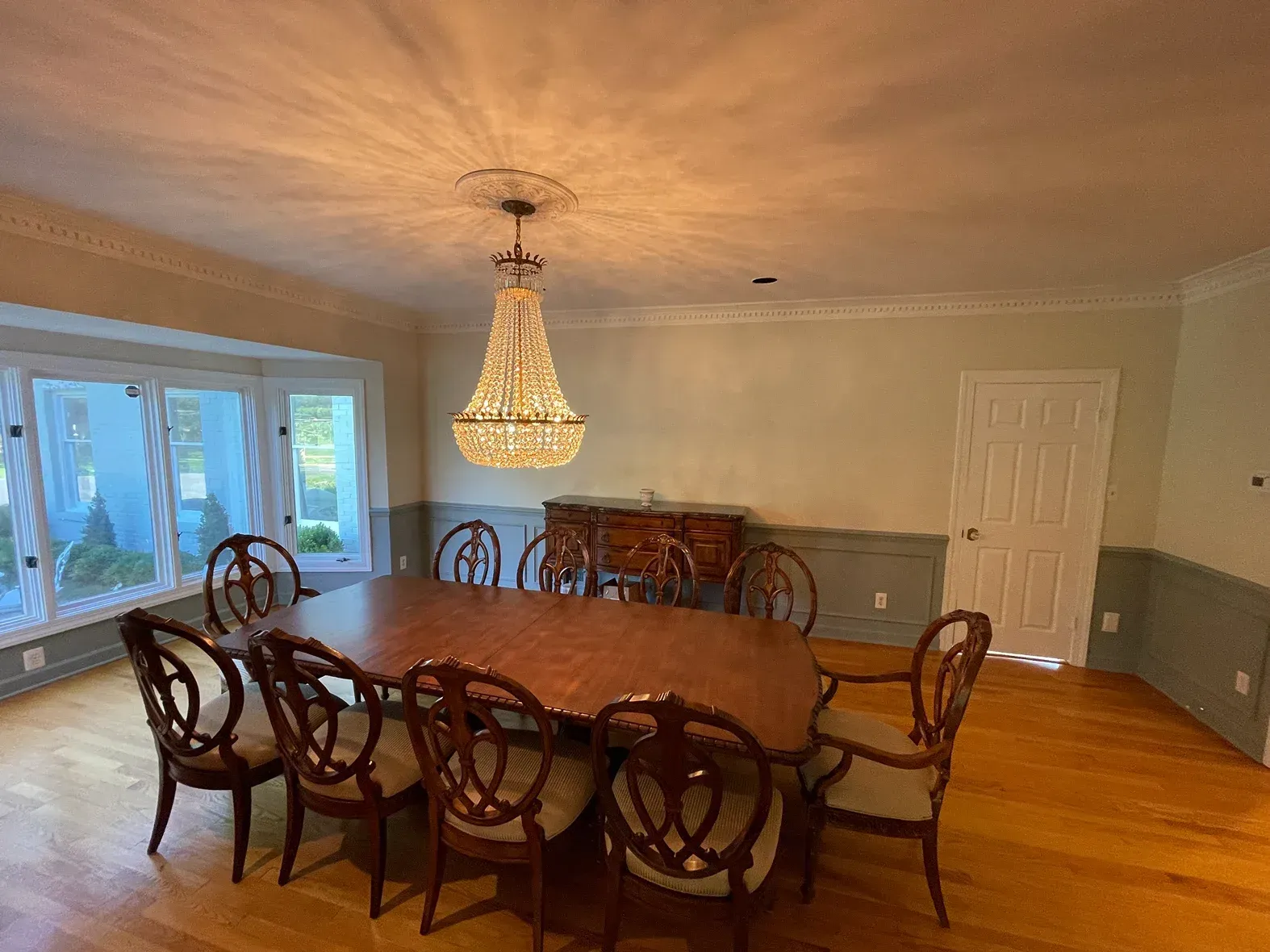 A dining room with a wooden table, chairs, a crystal chandelier, light-colored walls with wainscoting, and wood flooring.