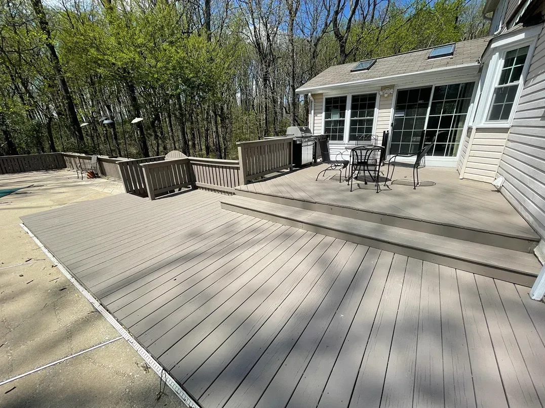 A multi-level light gray composite deck attached to a house, featuring a dining set, grill, and surrounding woods.