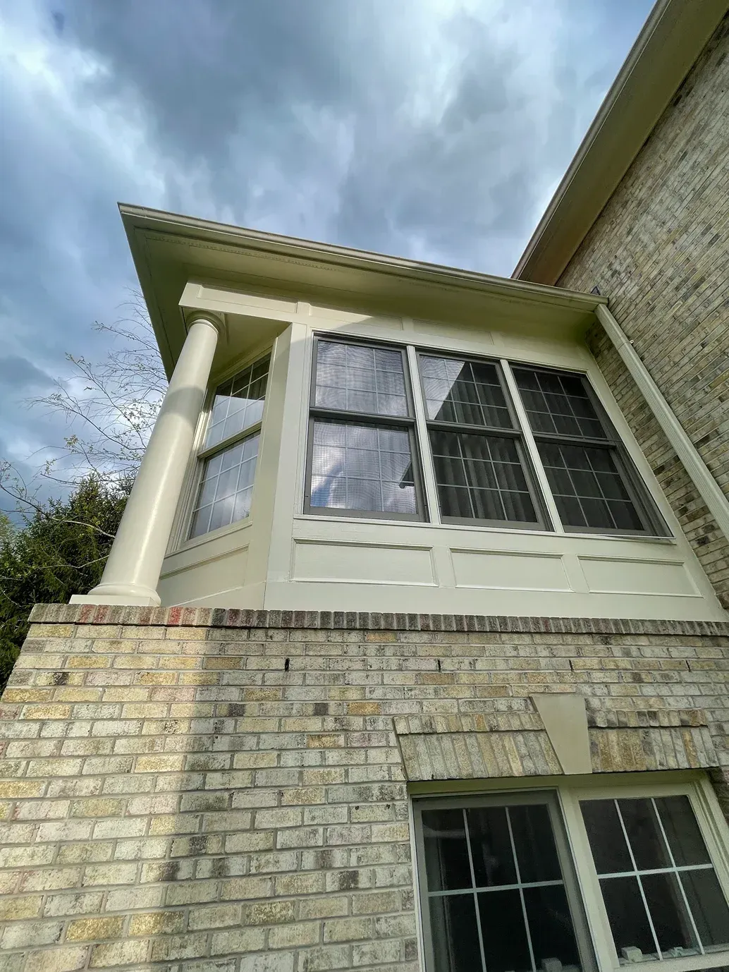 Low-angle view of a cream-colored sunroom addition with a white column, attached to a light brick house under a cloudy sky.