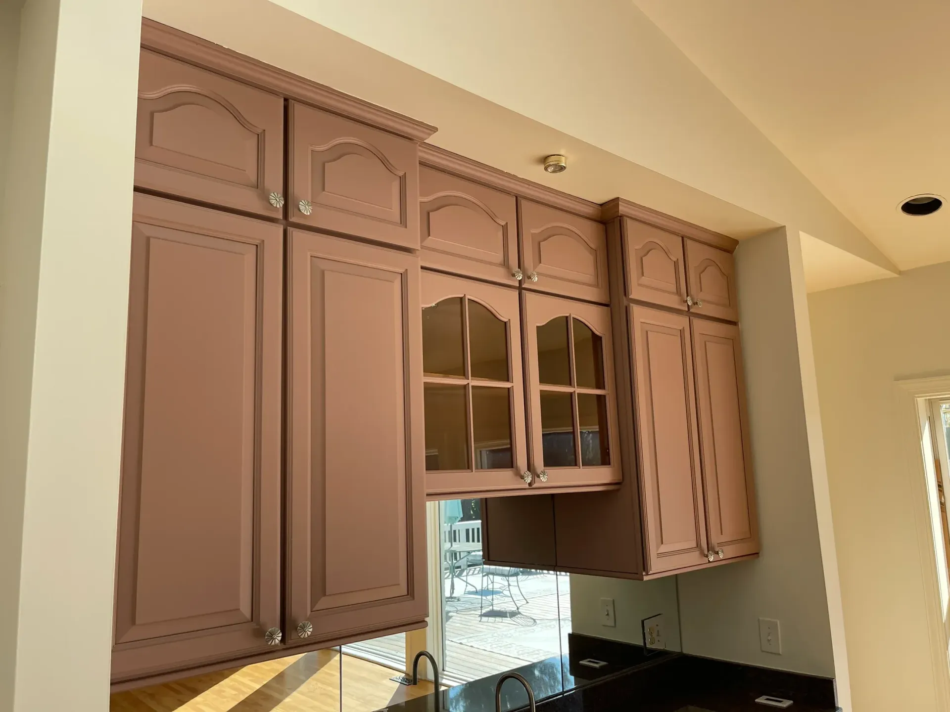 Dusty pink kitchen cabinets with arched doors and glass-fronted panels mounted above a counter near a window.