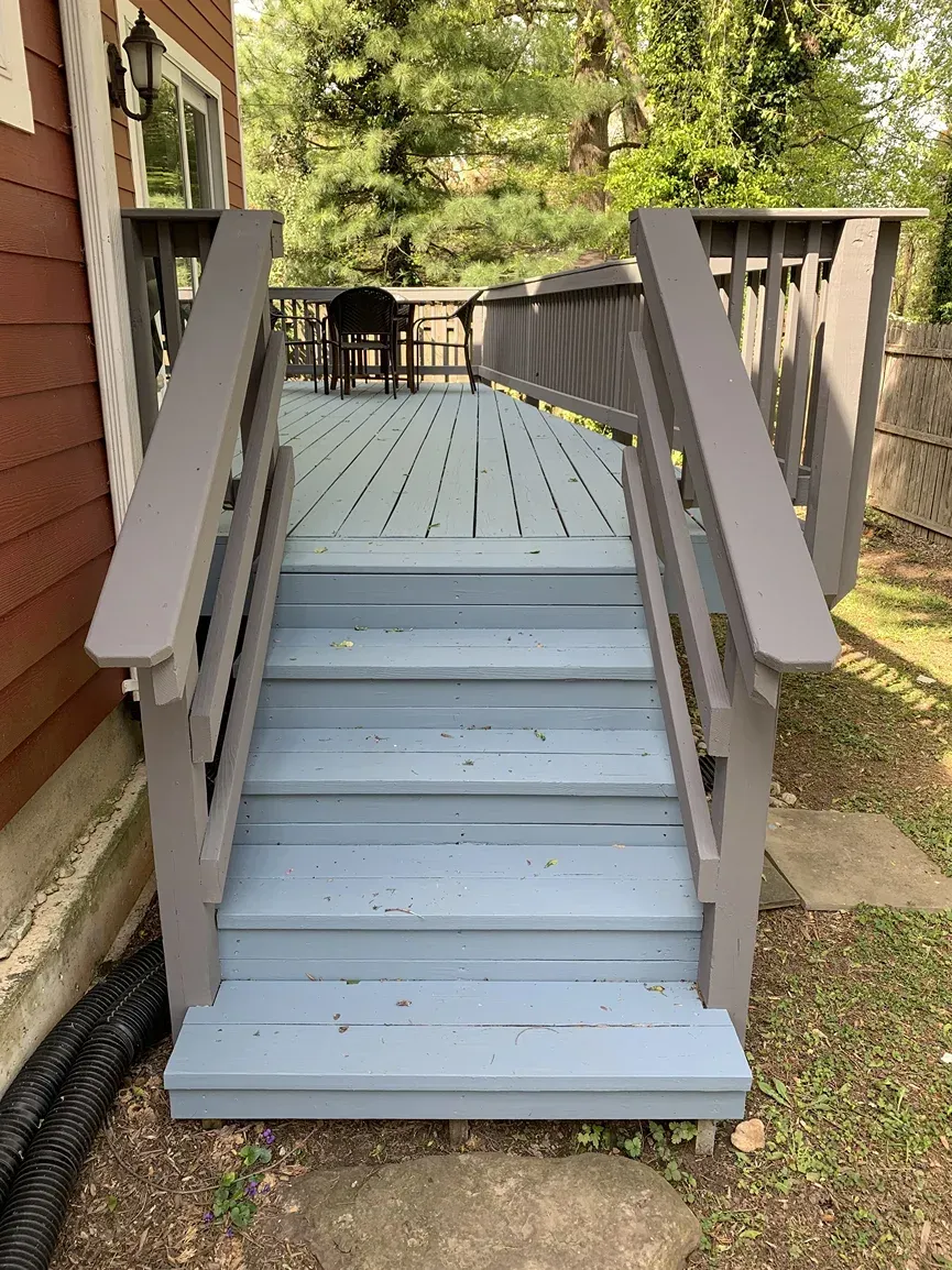 Light blue wooden deck stairs with grey railings leading up to a raised deck with patio furniture in a grassy yard.