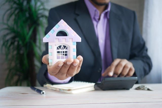 Person holding a house figurine while using a calculator