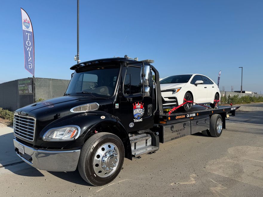 Black flatbed tow truck carrying a white car on a roadside under a clear blue sky