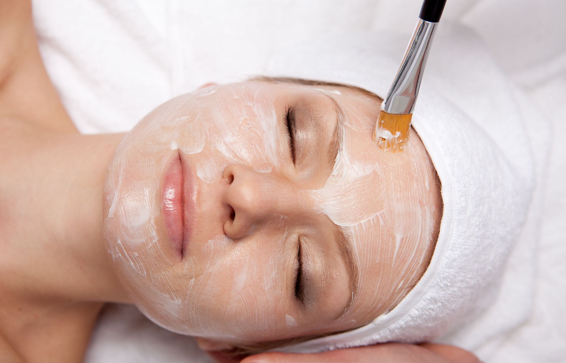 Woman receiving facial treatment, with cream on face, being applied by a brush.