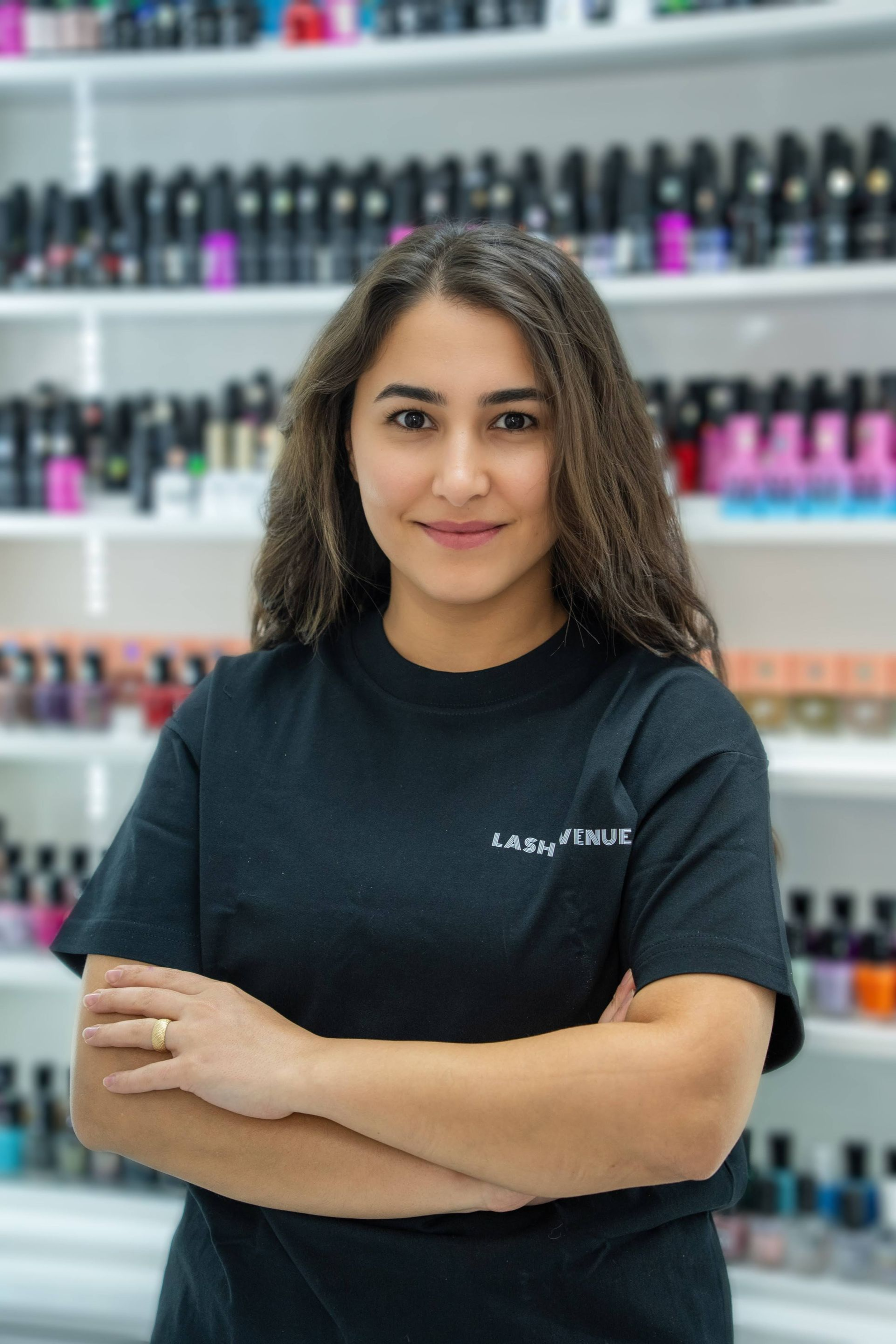 Woman in black shirt with arms crossed, smiling in a nail salon setting. Shelves of nail polish in background.