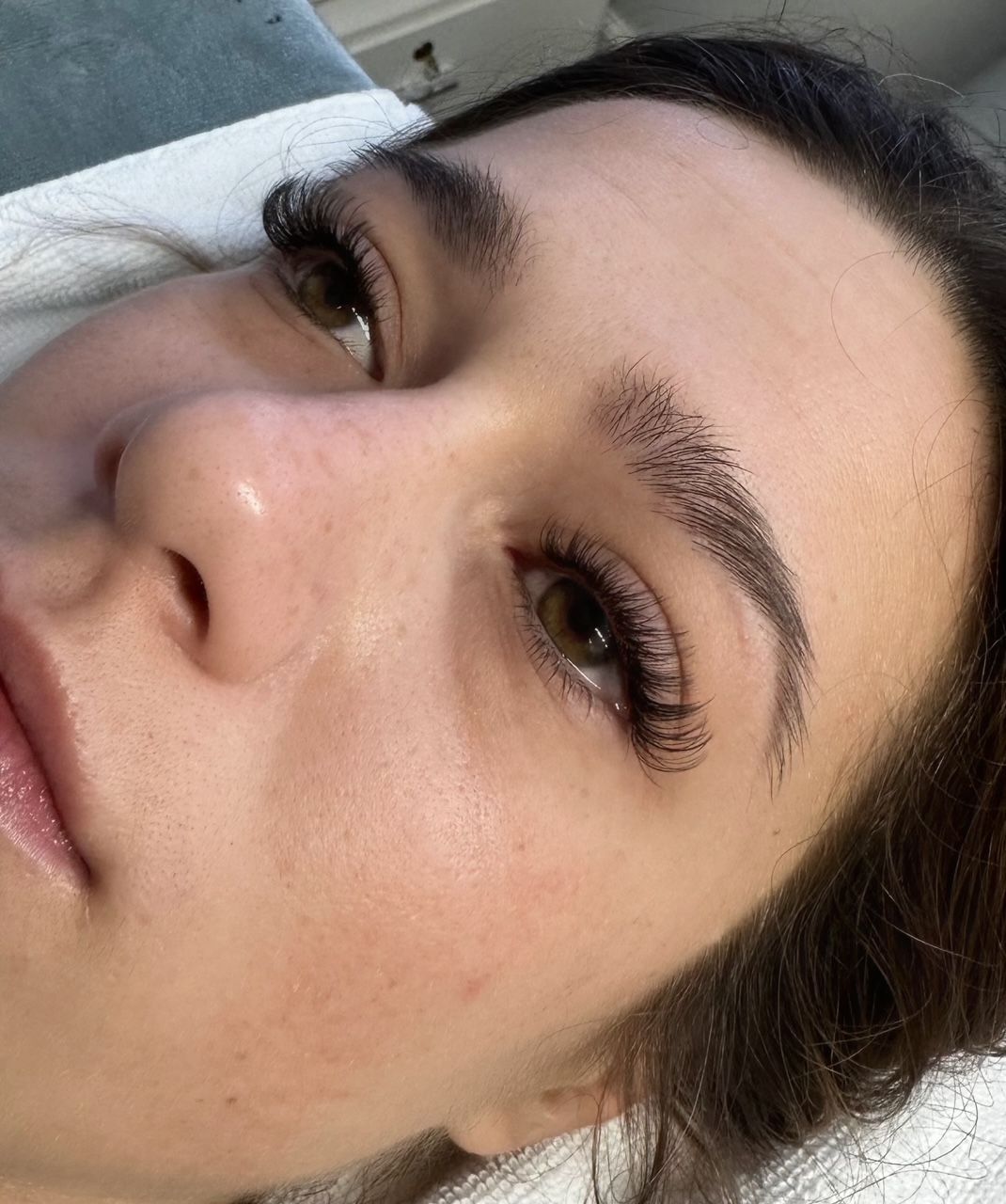 Woman's face close-up, eyes open, new microbladed eyebrow and lash extensions visible, in a medical setting.
