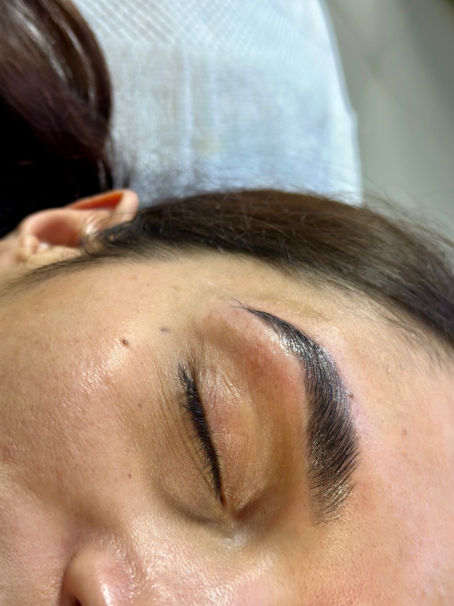 Close-up of a person's closed eye and eyebrow after a cosmetic procedure. The eyebrow is dark and appears freshly tattooed.