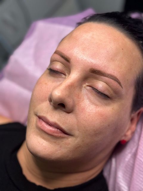 Woman with closed eyes, newly tattooed brown eyebrows, resting on a pink sheet.