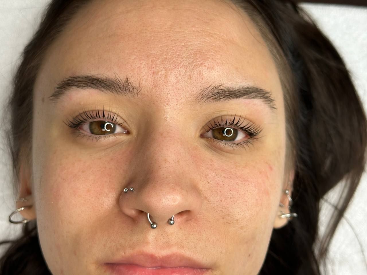 Woman with brown eyes and multiple facial piercings, post-eyebrow and eyelash procedure.