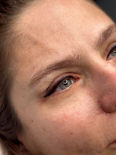Close-up of a person's eye with eyeliner and brow tattoo. The skin is fair with light brown brows.