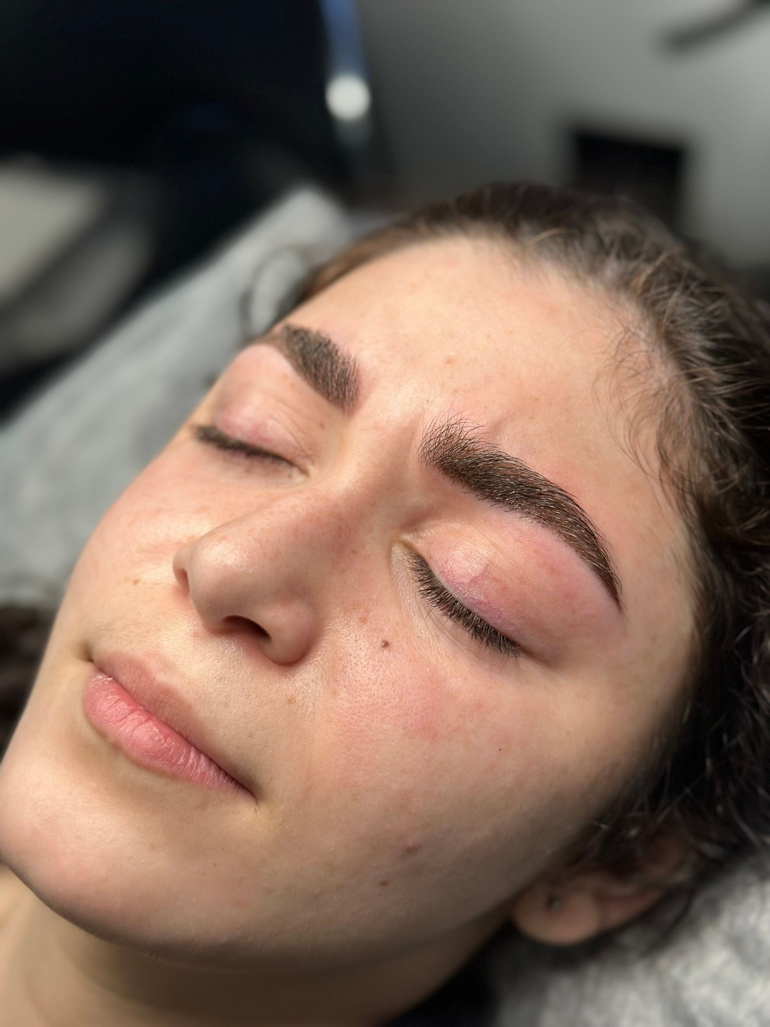 Woman with closed eyes, newly tattooed eyebrows. Face resting on a pillow, skin appears irritated with slight redness.
