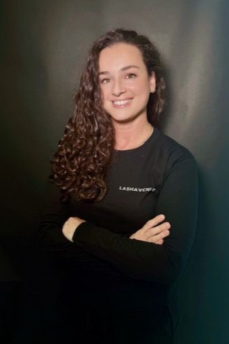 Woman with short brown hair smiles, wearing a black long-sleeved shirt, leaning against a white wall.