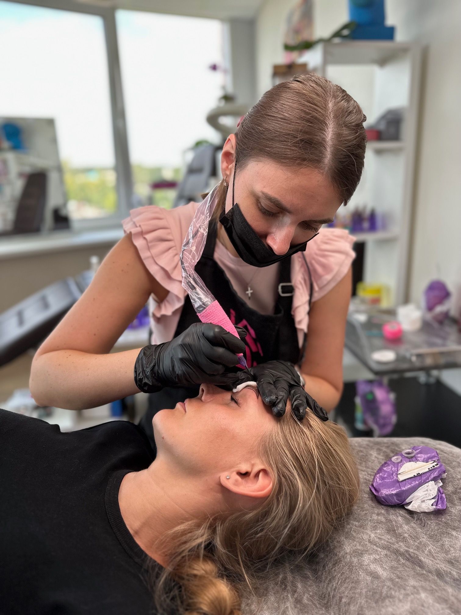Person getting eyebrow microblading at a salon. Technician wears a mask, gloves, and apron.