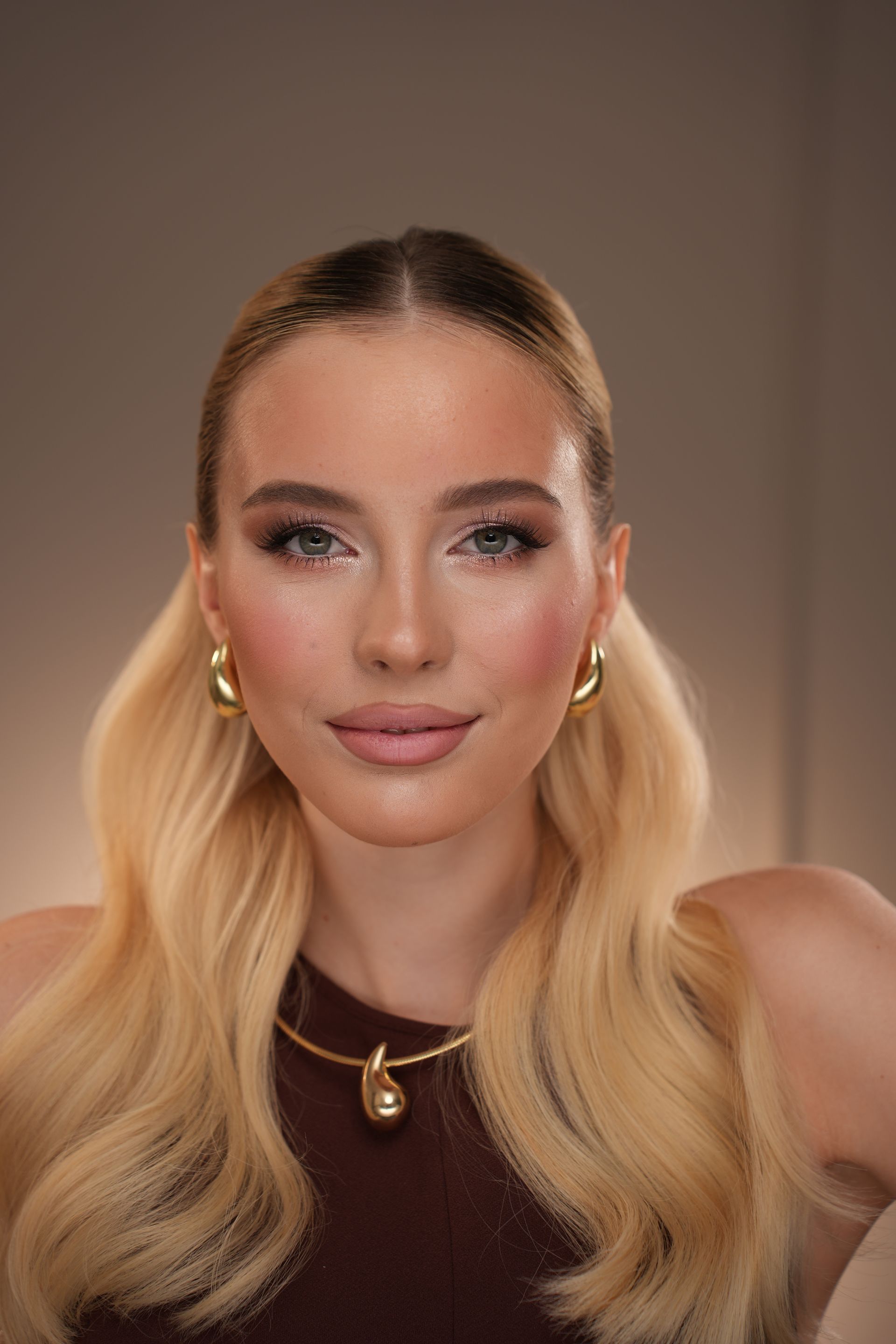 Blonde woman with wavy hair, makeup, wearing gold jewelry, brown top, and smiling at the camera.